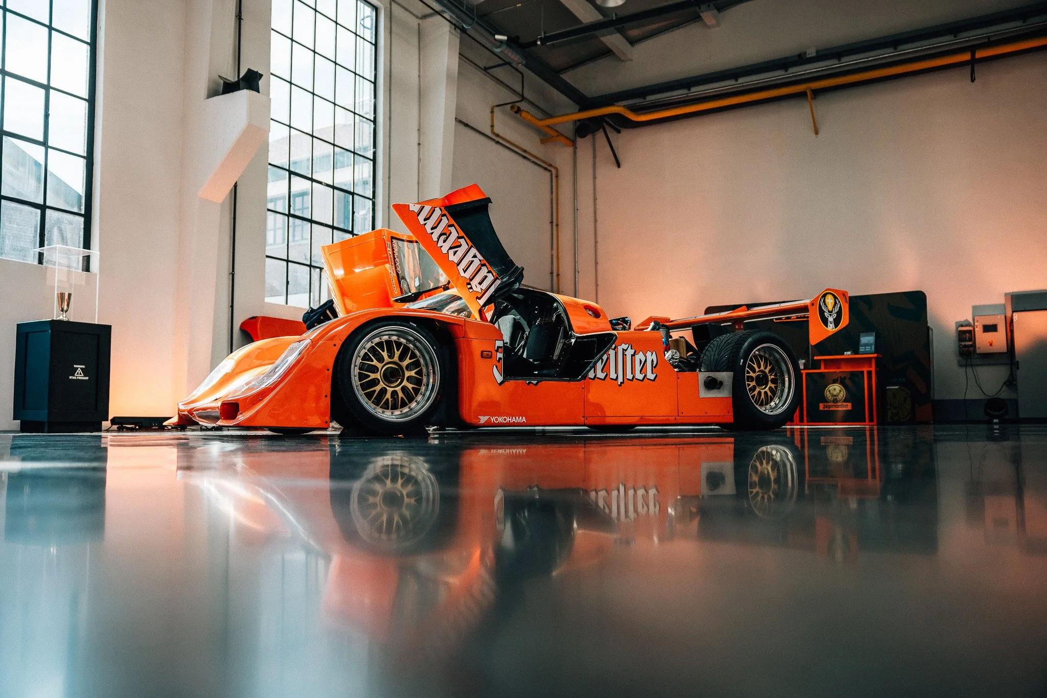 An orange race car with sponsor logos displayed indoors, with a polished reflective floor and large windows in the background.
