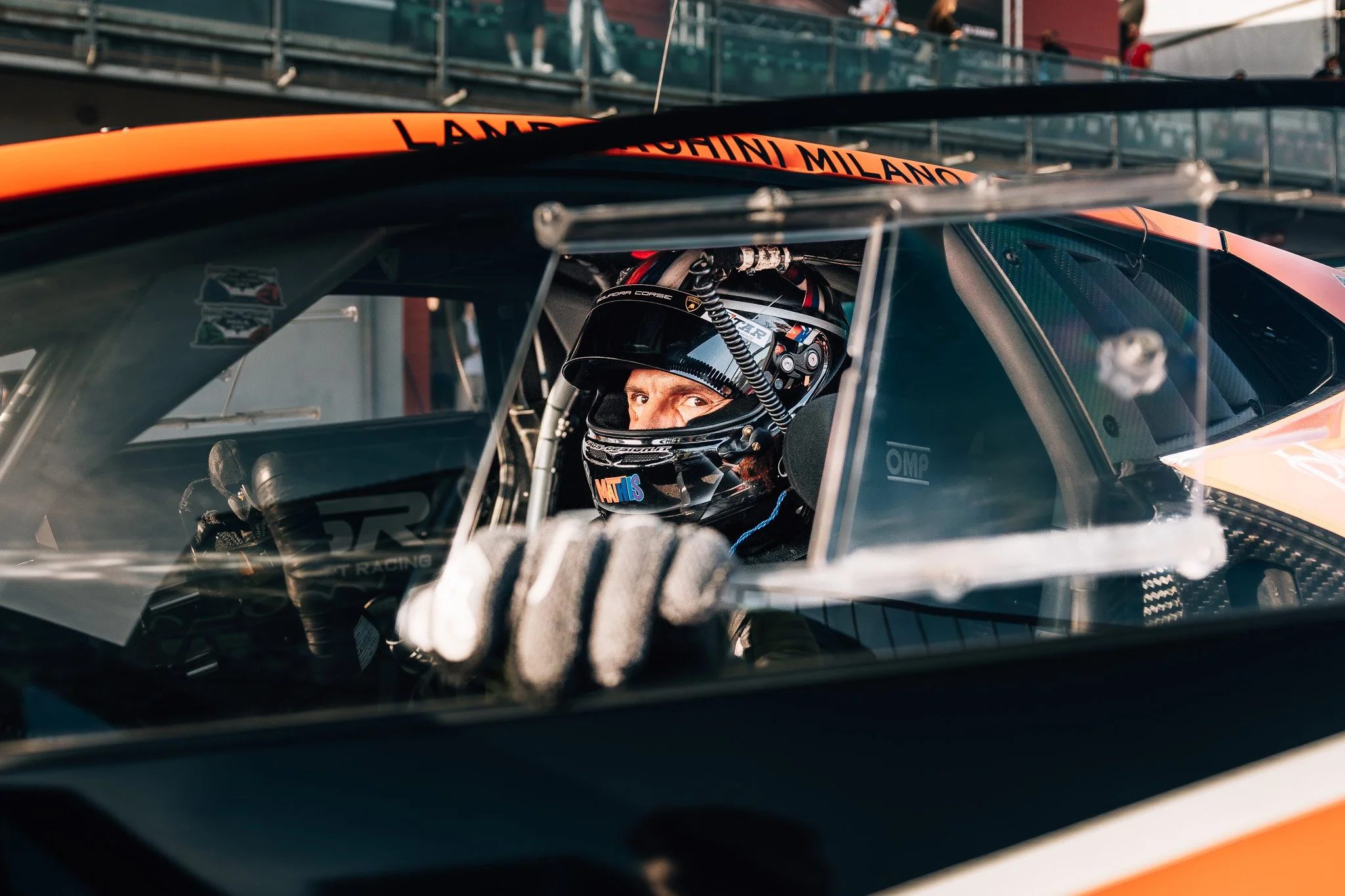 Race car driver wearing a helmet, sitting inside a race car, looking out of the window with hand on the steering wheel, with the race track and spectators in the background.