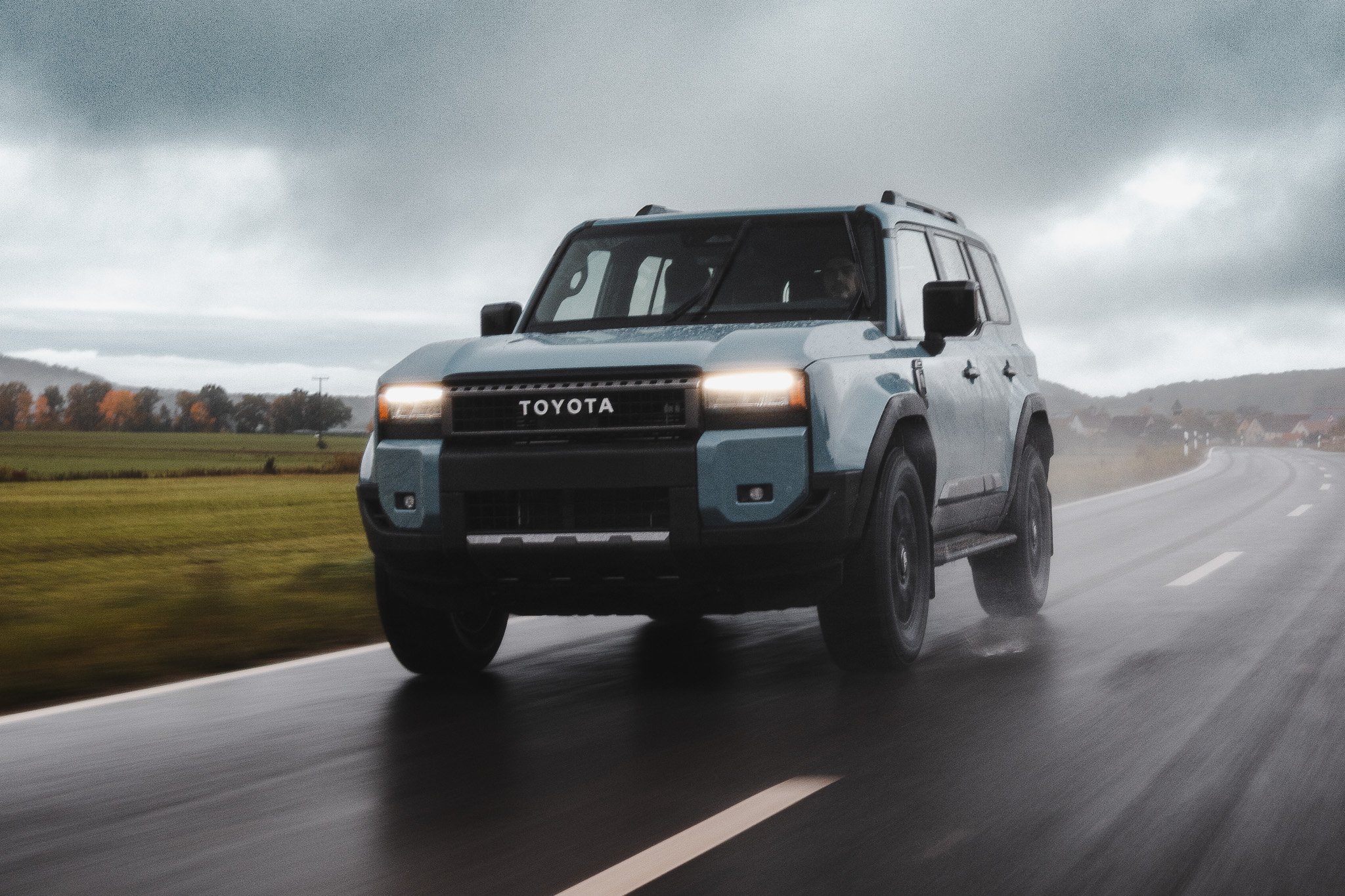 A light blue Toyota SUV driving on a wet road during cloudy weather with fields and trees in the background.