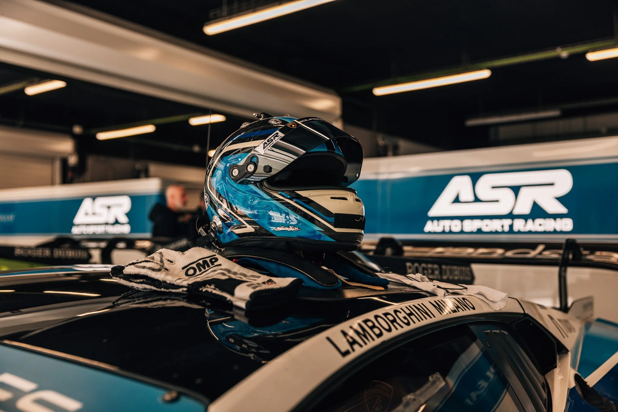 A racing helmet and racing gloves placed on the roof of a race car, with a blurred background of a garage or paddock area displaying a large sign that reads 'ASTR AUTO SPORT RACING'.