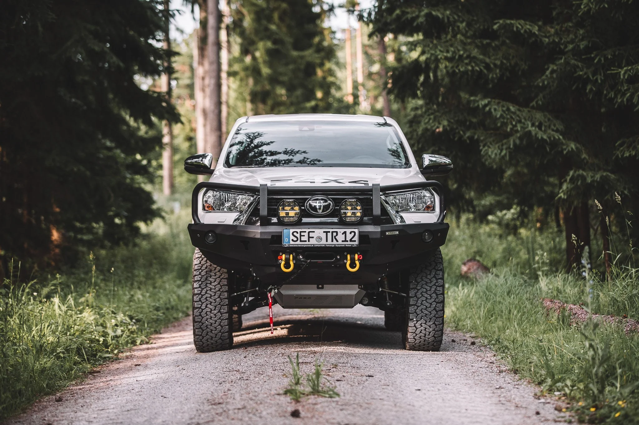 A white off-road vehicle with a black bumper guard and large tires parked on a forest trail surrounded by tall green trees.