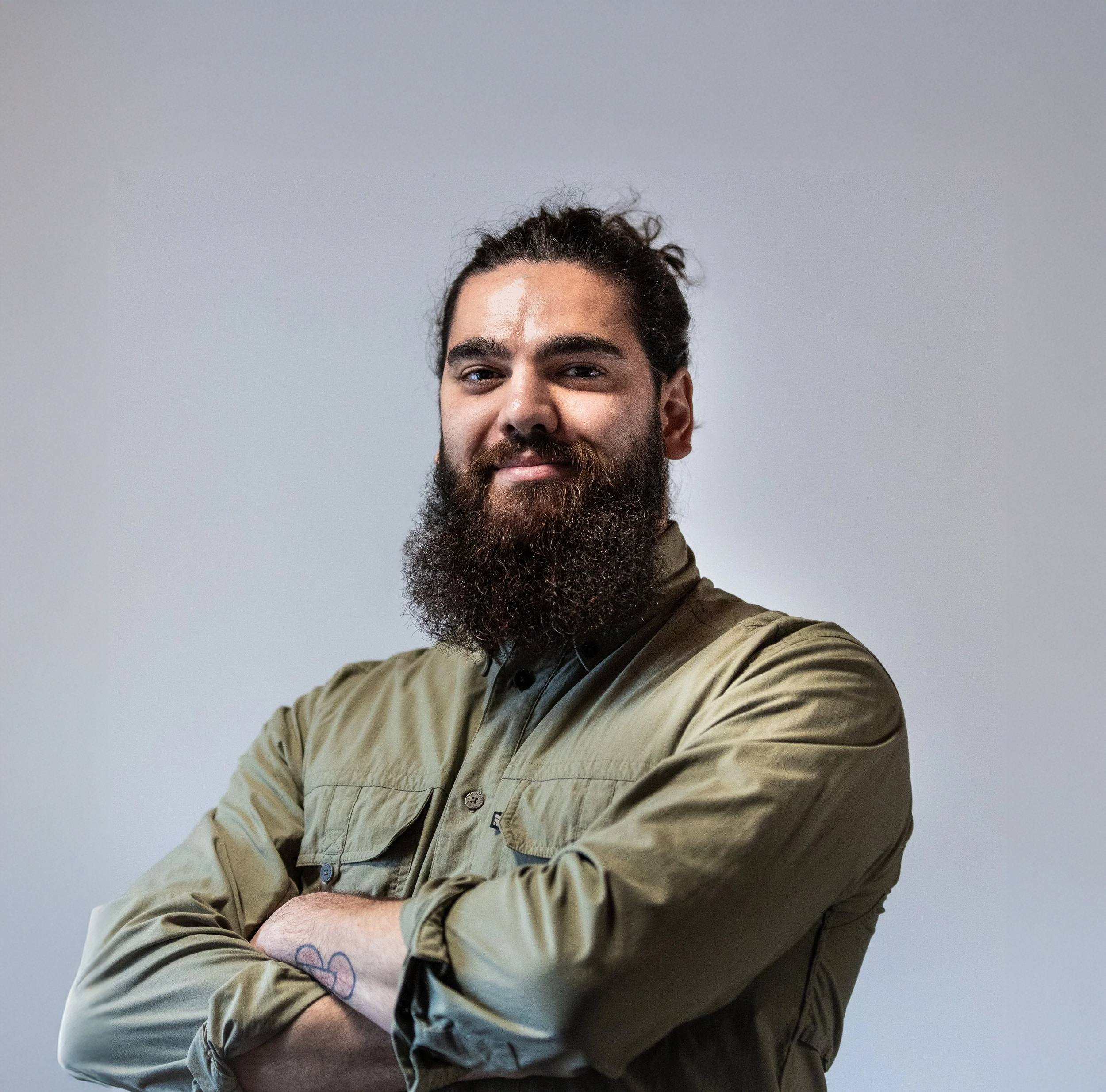 A bearded man with dark curly hair smiling, wearing a khaki jacket with rolled-up sleeves, standing against a plain gray background.