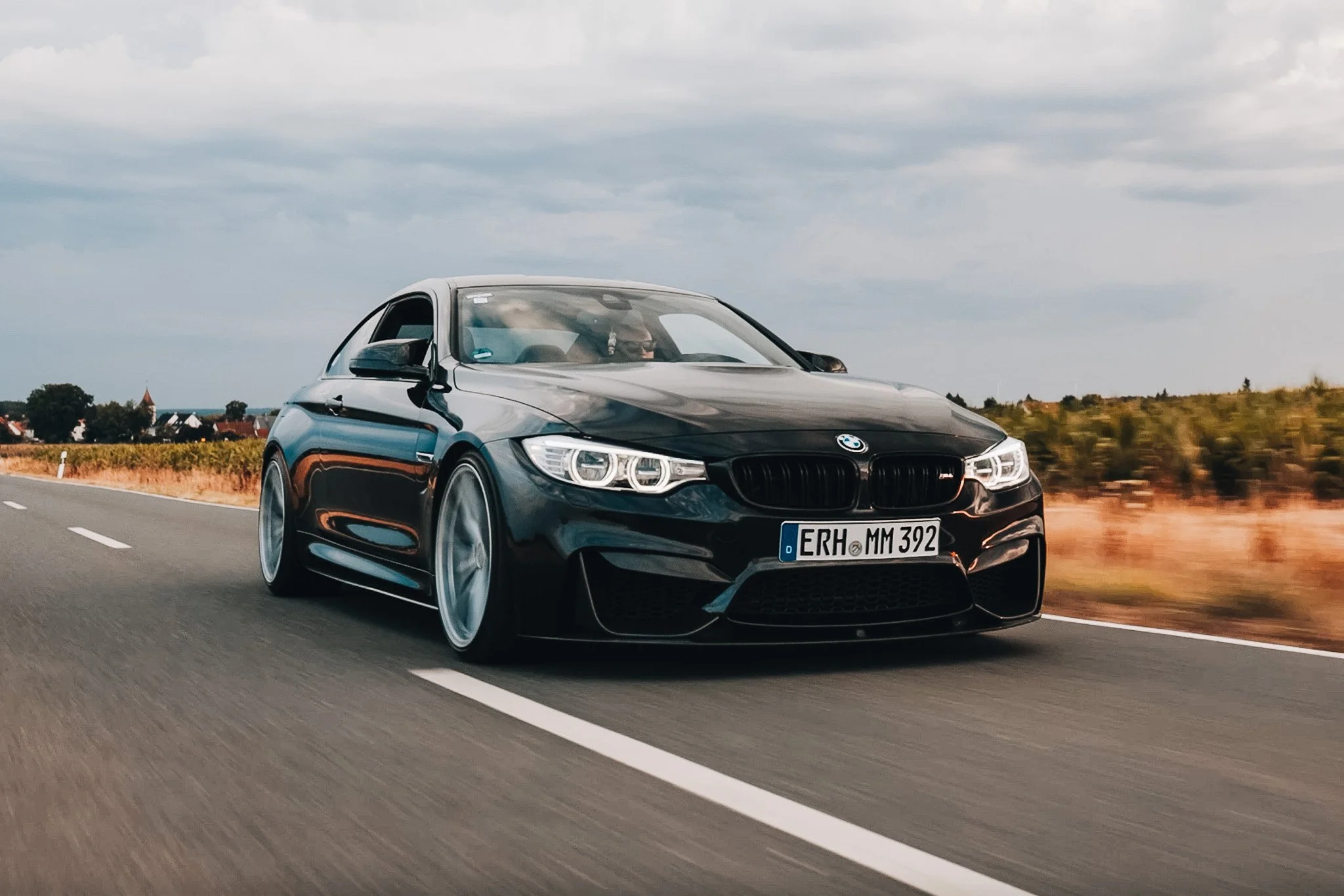 A black BMW car driving on a highway with a blurred background of fields and houses under a cloudy sky.