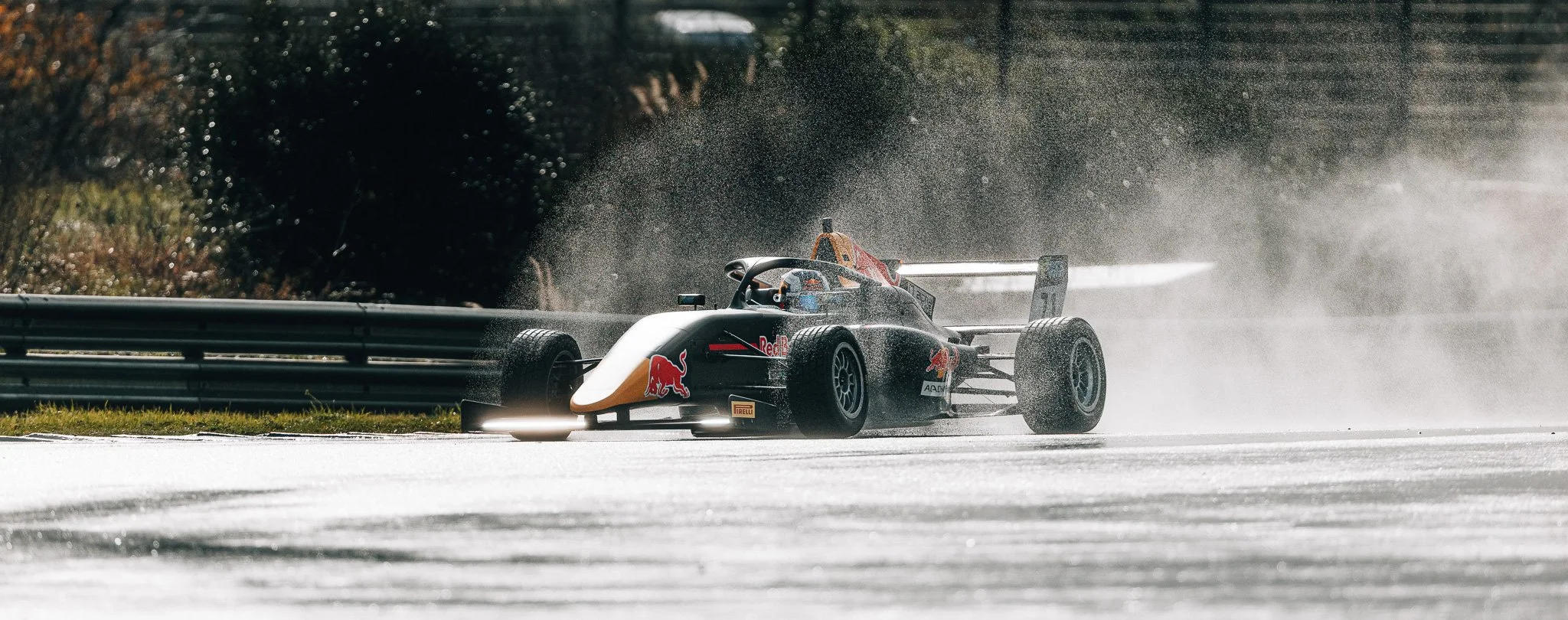A race car driving on a wet track, creating splashes of water as it speeds by, with a backdrop of trees and a metal guardrail.