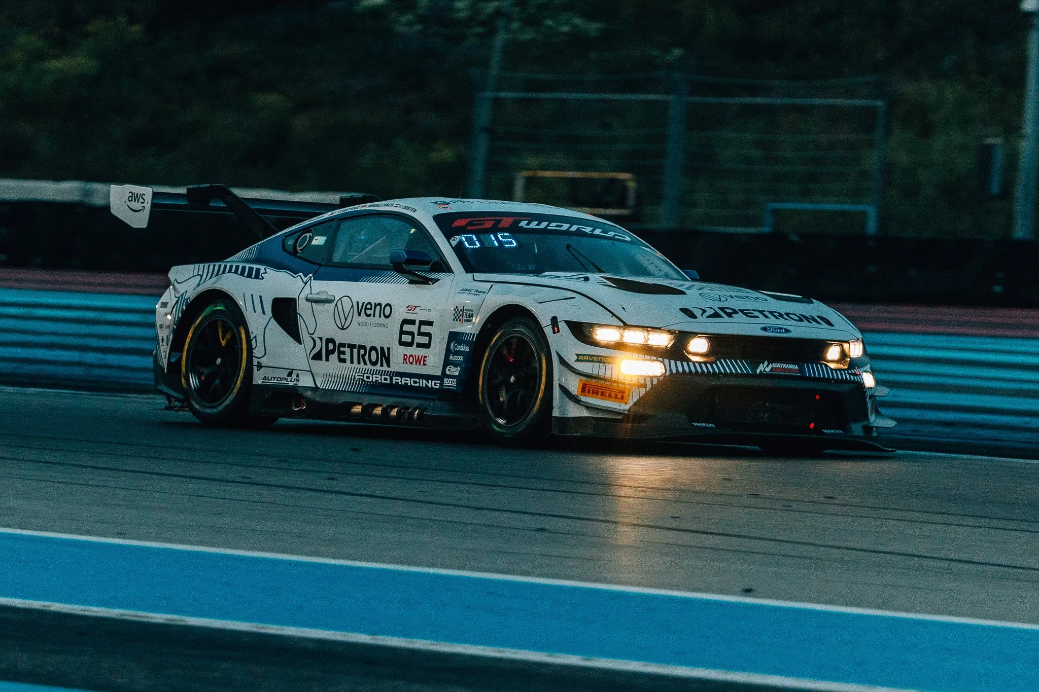 A white race car with sponsor logos, including Petron and Veno, speeding on a race track at night with its headlights on, surrounded by a dark background with trees.