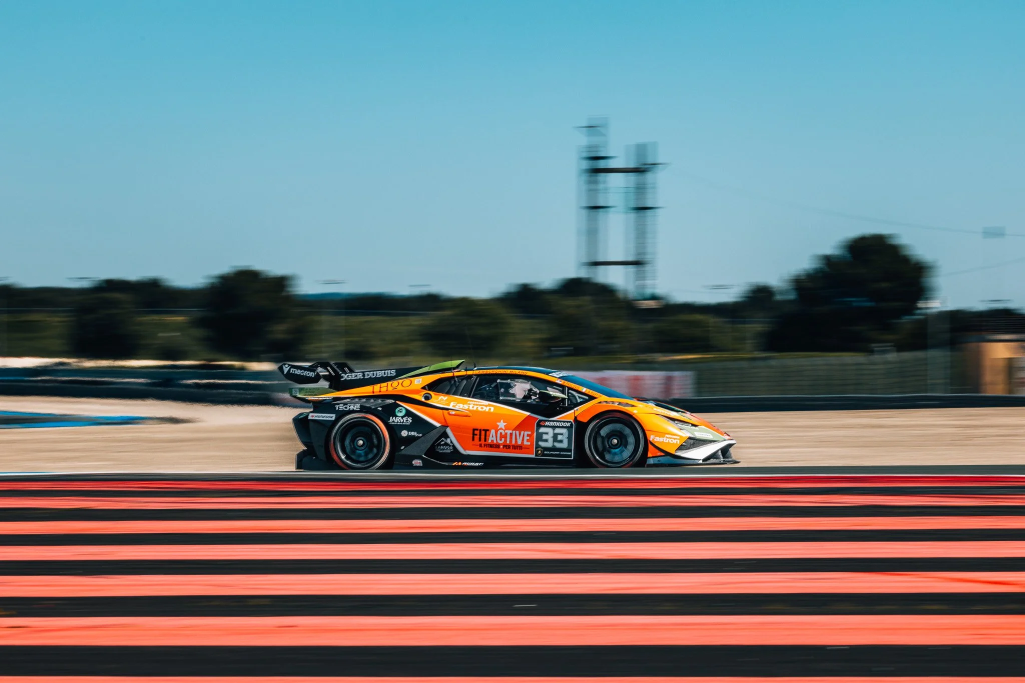 An orange and black race car moving at high speed on a race track during the daytime with a blue sky and trees in the background.