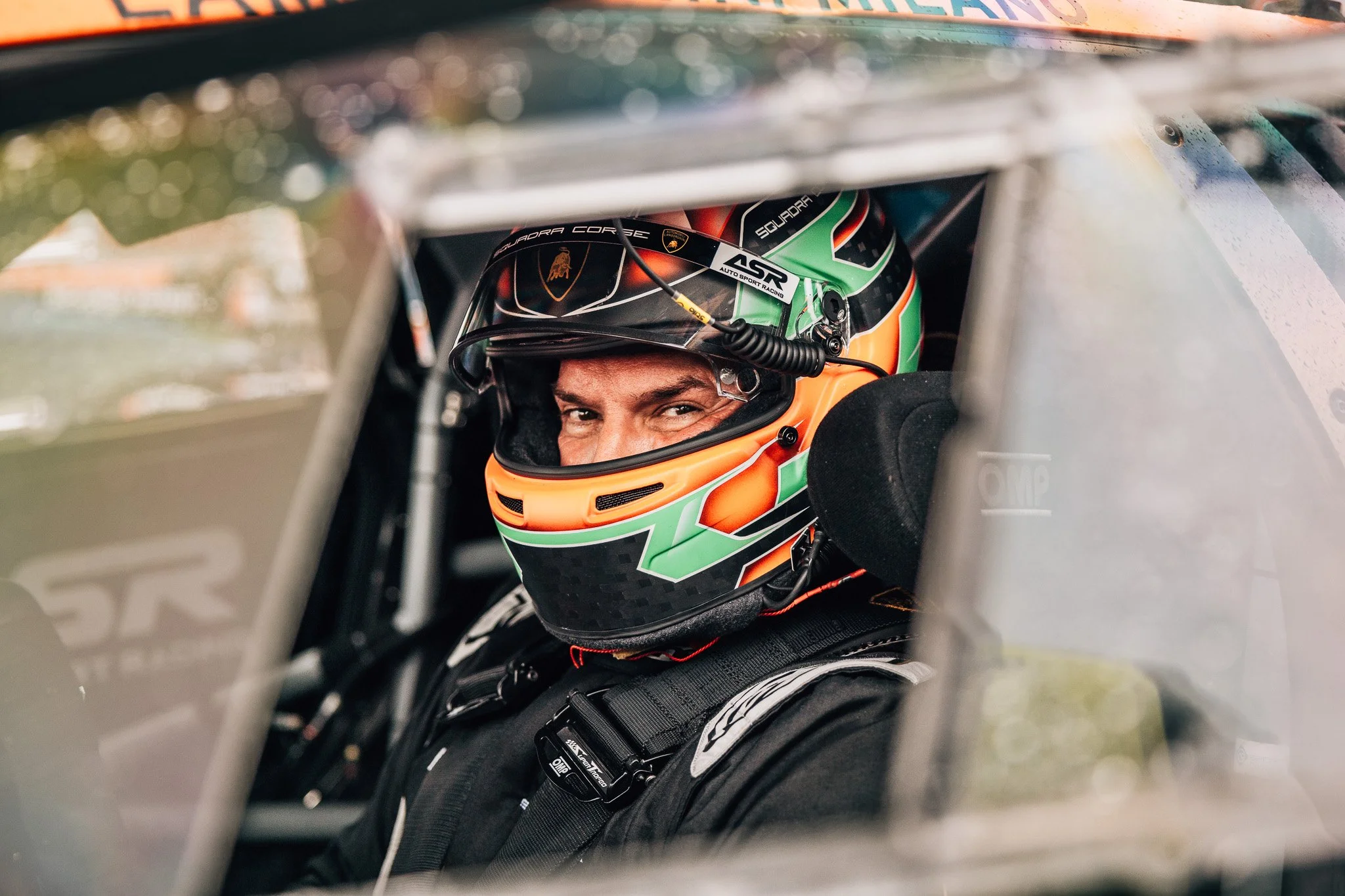 Close-up of a race car driver wearing a colorful helmet with green, orange, and black design. The driver is seated inside the cockpit of a race car, looking out through the opening, with a smile visible on their face.