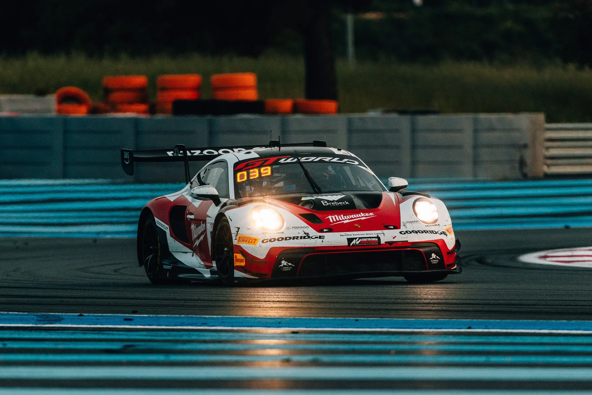 A red, white, and black race car on a track at dusk, with blue barriers in the foreground and orange and black tires stacked in the background.