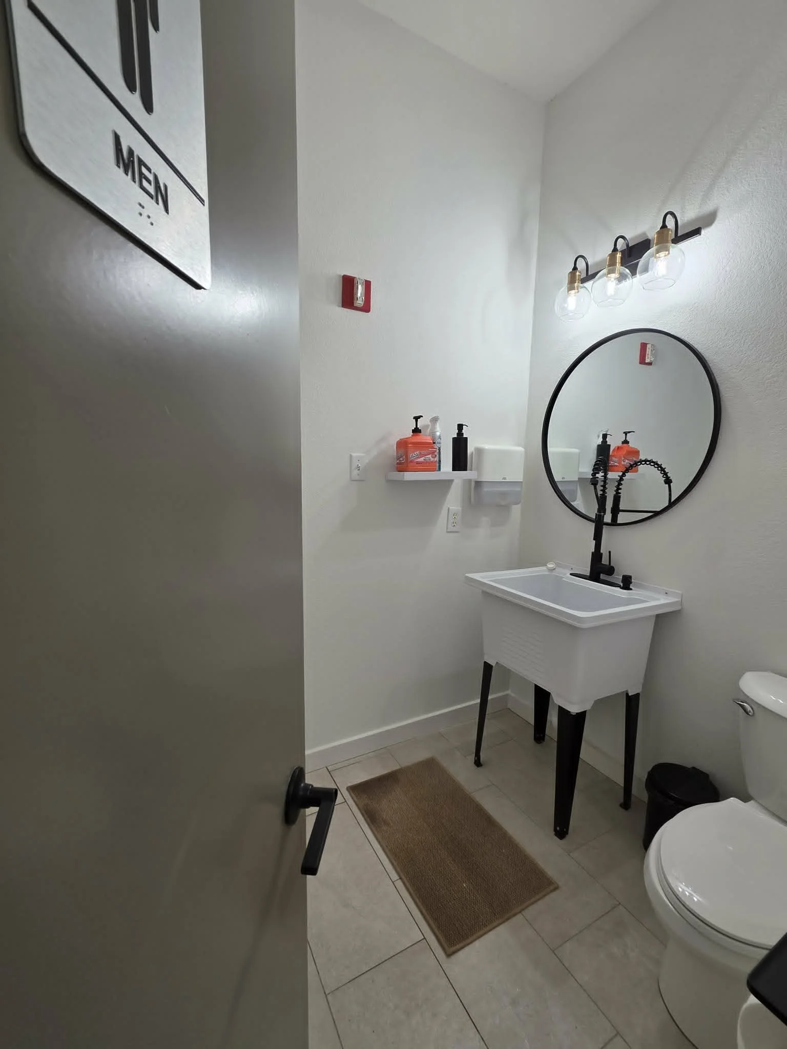 A clean restroom with white walls, a white utility sink, a round mirror, a wall-mounted light fixture with three glass shades, a small shelf holding hand soap and sanitizer, a brown rectangular rug, and a toilet visible in the bottom right corner.