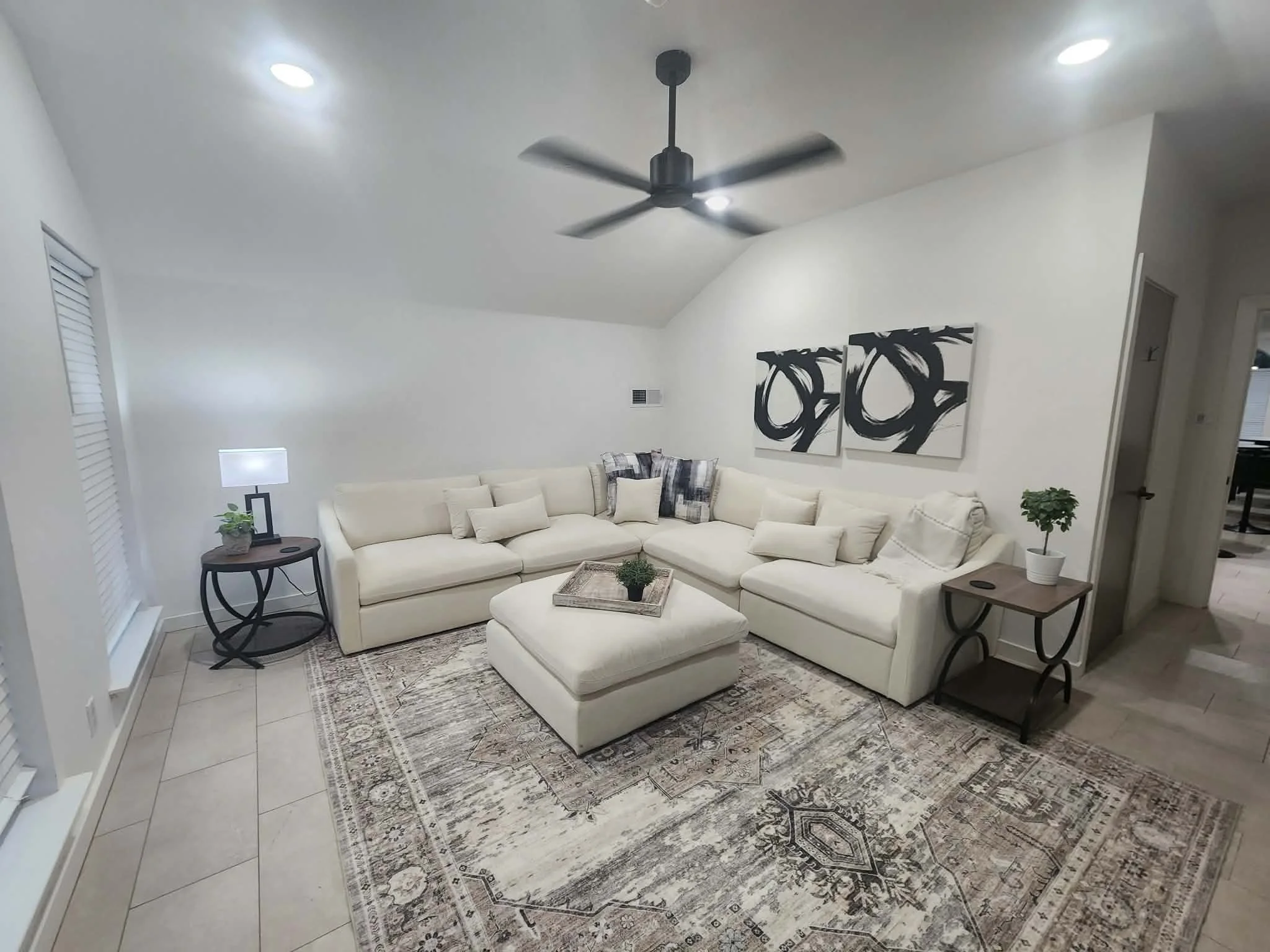 Living room with white sectional sofa, a rectangular coffee table, a patterned area rug, two matching black side tables with plants, a lamp, and wall art with black abstract designs.