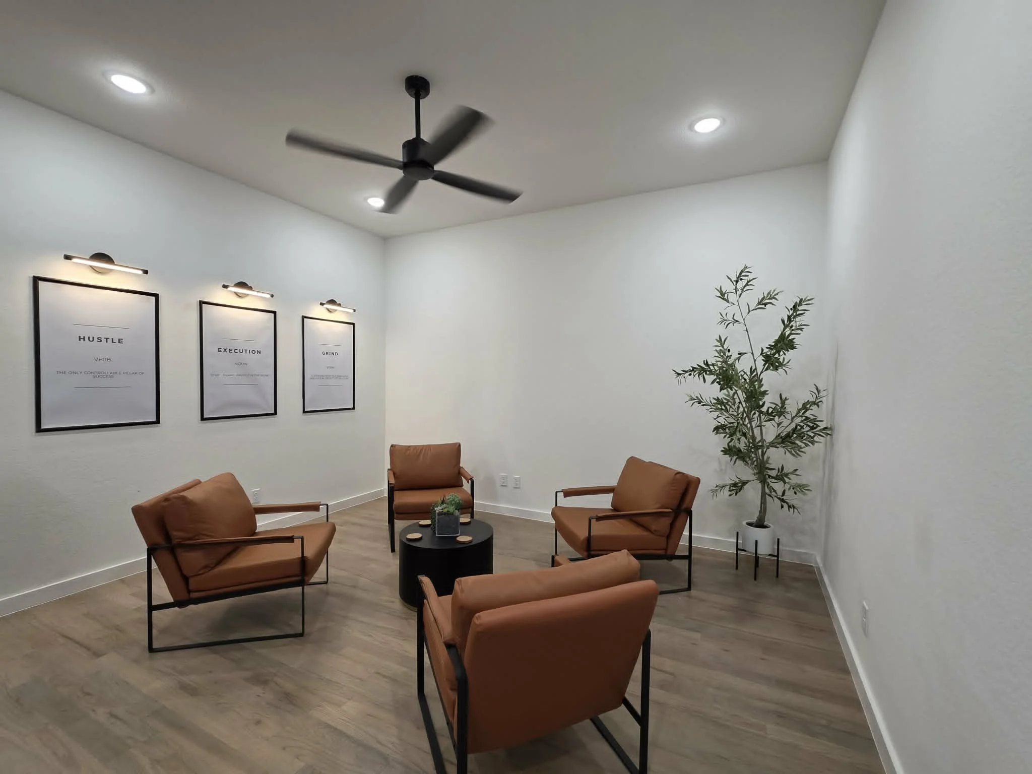 A modern waiting room with four brown armchairs arranged around a small round black table, a tall green plant in a white pot, and framed motivational posters on the white wall. The ceiling has recessed lighting and a black ceiling fan.