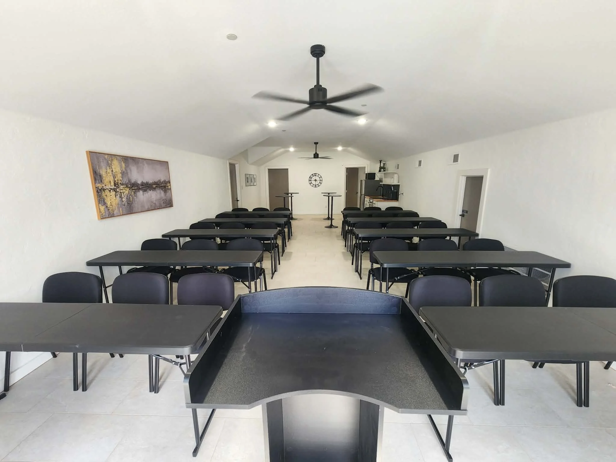 Conference room with black tables and chairs, white walls, ceiling fans, framed artwork, and a wall clock.