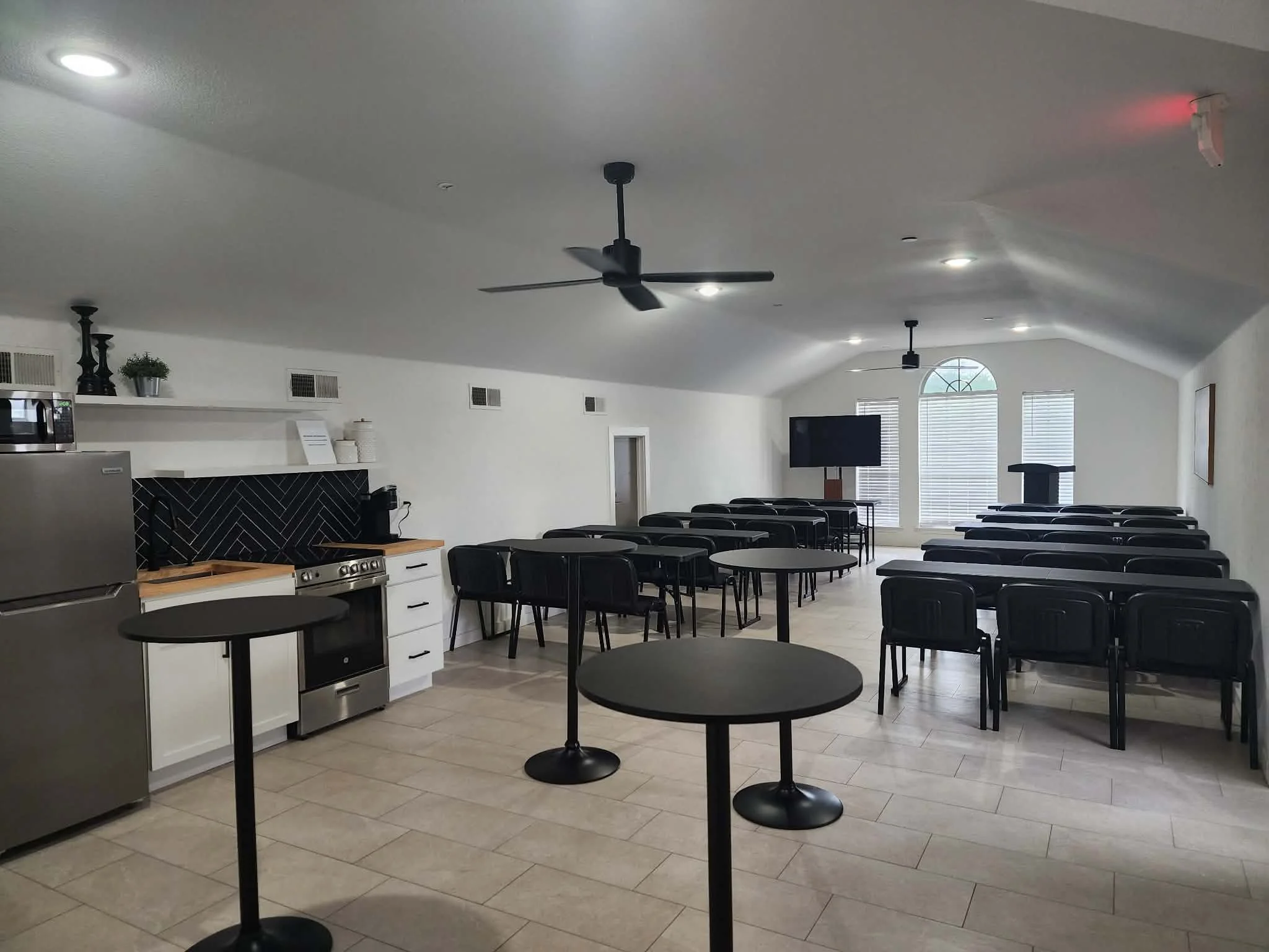 Empty room with black round tables and chairs, white kitchen cabinets, stainless steel appliances, ceiling fans, a television, and window blinds.