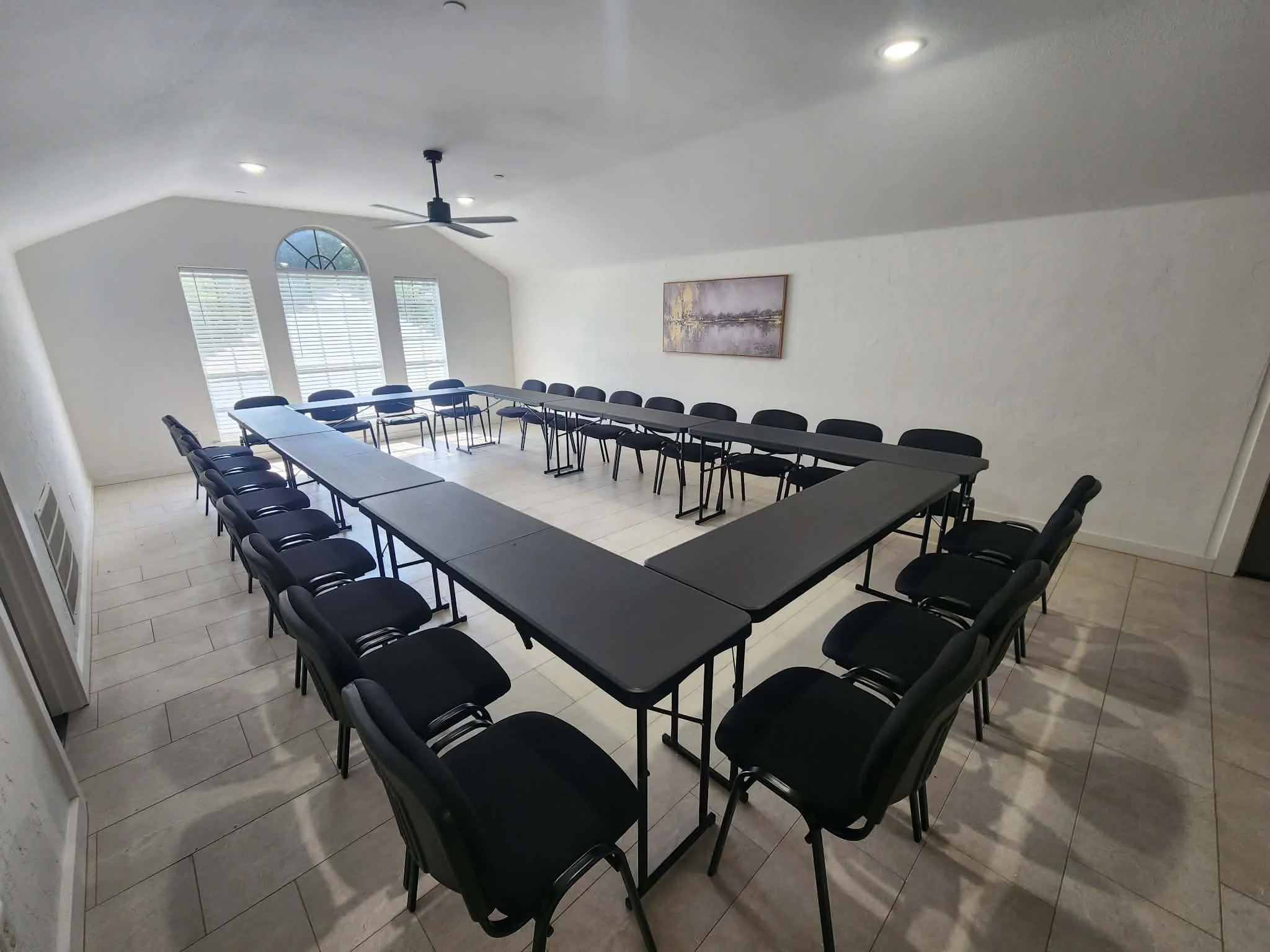 A conference room with a U-shaped arrangement of black chairs and gray tables. There are three windows with blinds, a ceiling fan, and a framed artwork on the wall depicting a landscape scene.