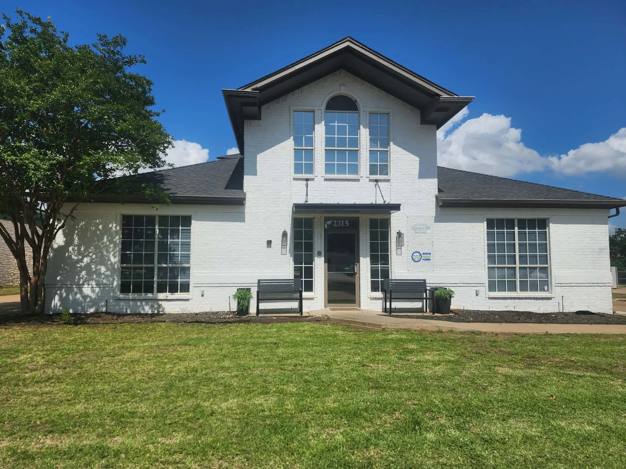 White two-story house with large front windows, a black front door, and a lawn with trees, under a blue sky with some clouds.