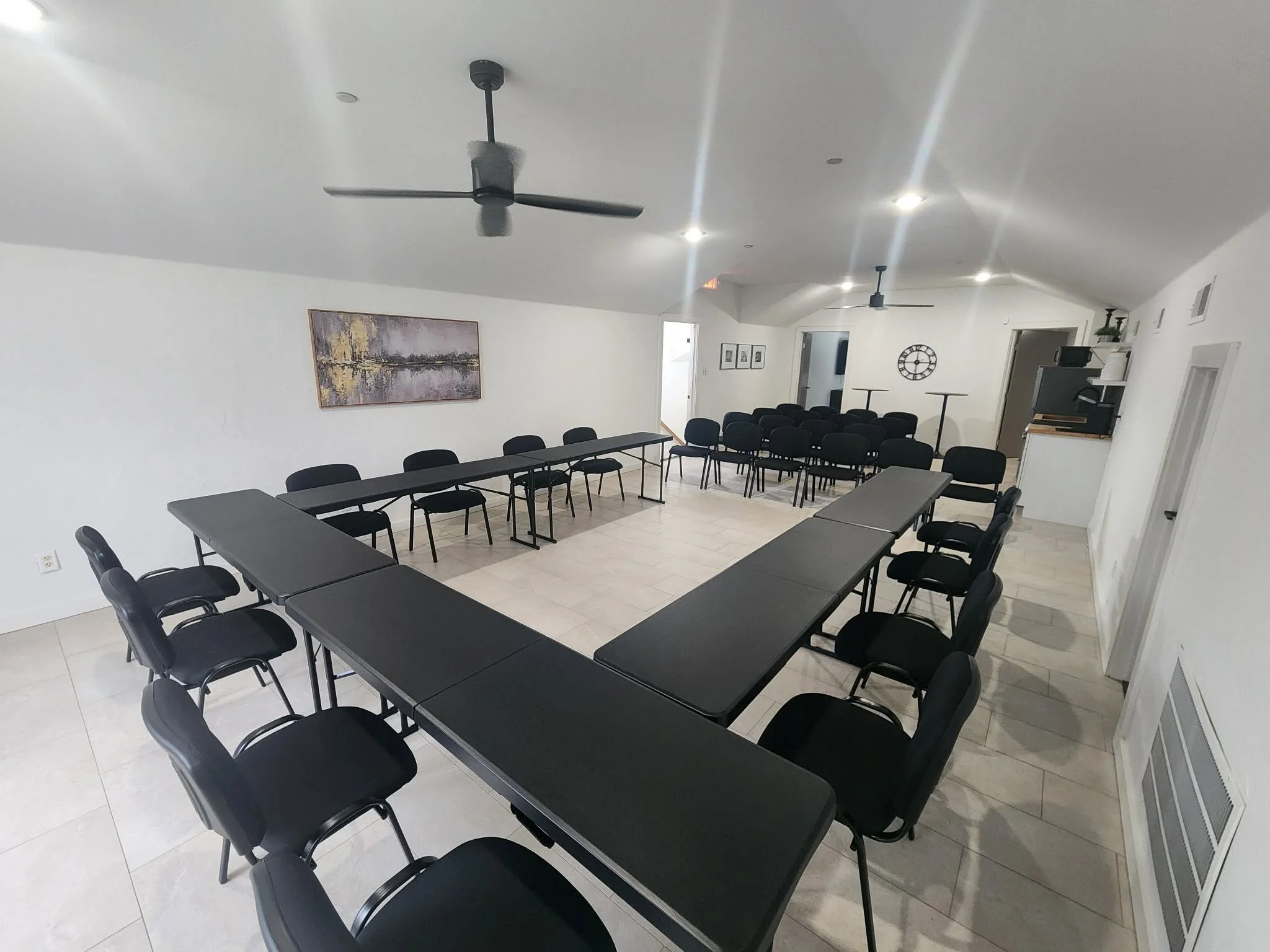 A spacious conference room with black tables and chairs arranged around the room. The room has white walls, ceiling fans, and a wall clock. There is artwork on the wall and a doorway leading to another room.