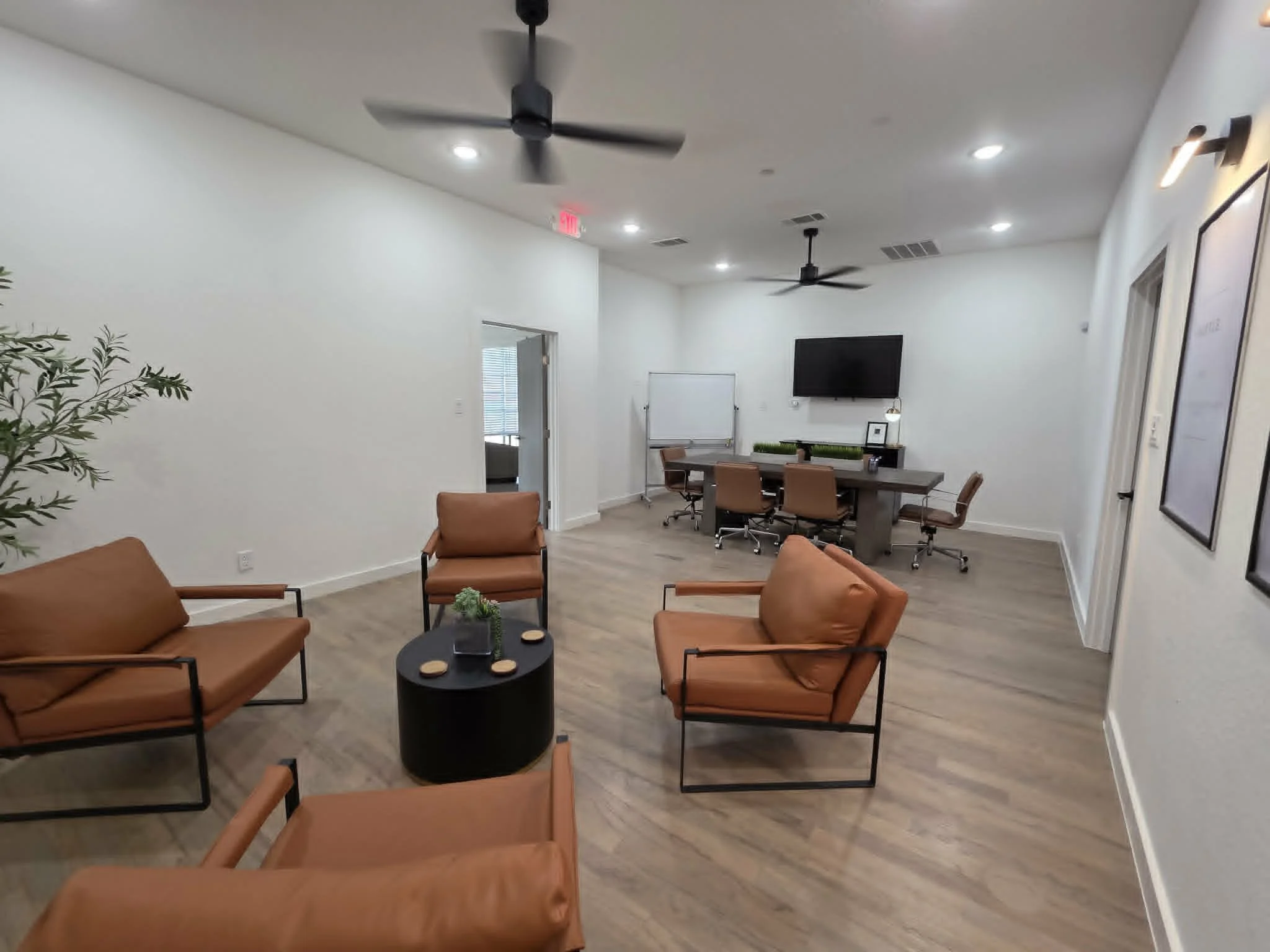 A modern conference or meeting room with white walls and ceiling, featuring three ceiling fans, multiple recessed lights, a wall-mounted flat-screen TV, and framed art on right wall. The room has a long table with chairs around it, a whiteboard at the back, and a small black round coffee table with decorative coasters and a small plant in the foreground.