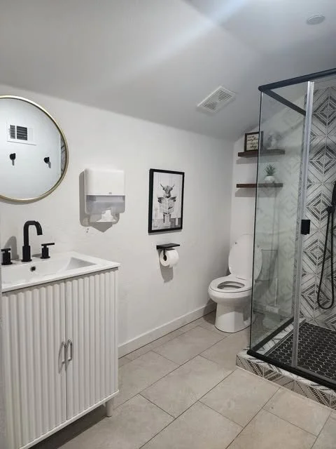 Modern bathroom with white walls, round mirror, black fixtures, a white sink with vertical slats cabinet, a framed artwork, a toilet, and a glass shower with patterned tiles.