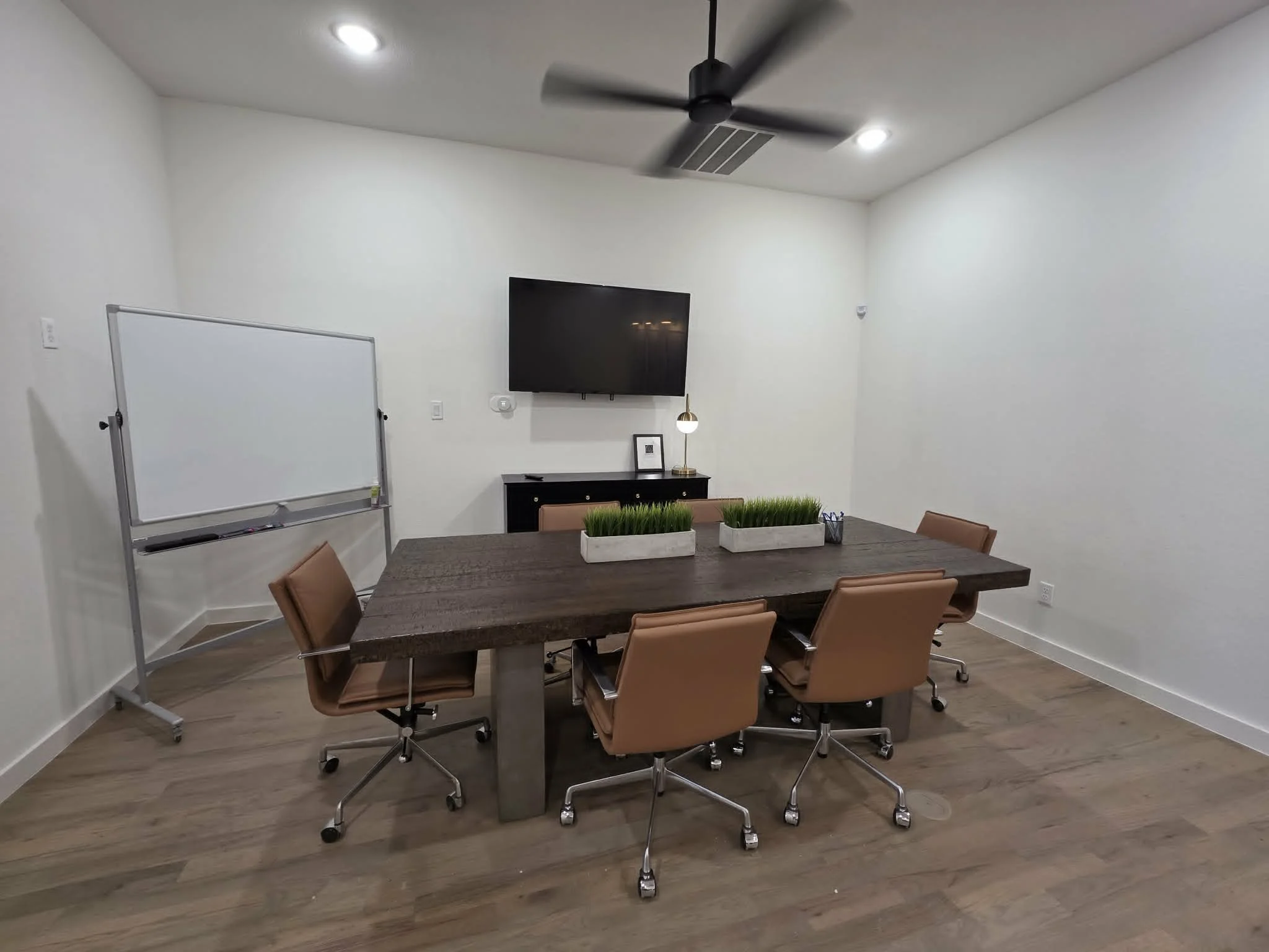 Meeting room with a wooden table, tan chairs, a whiteboard, a wall-mounted TV, and two planters.