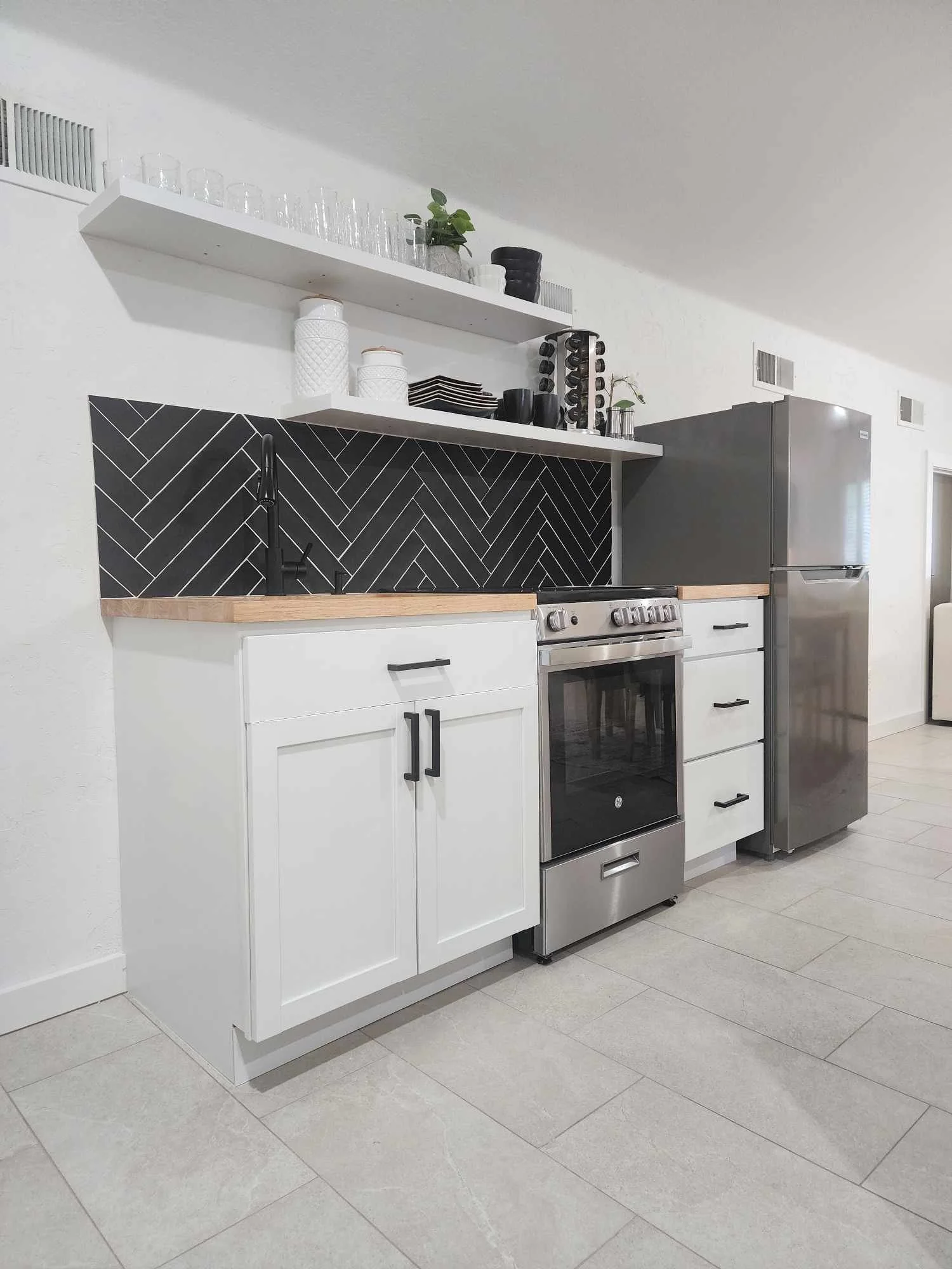 Modern kitchen with white cabinets, black and wood accents, and stainless steel appliances, including a refrigerator and oven, with open shelving and black dishware.