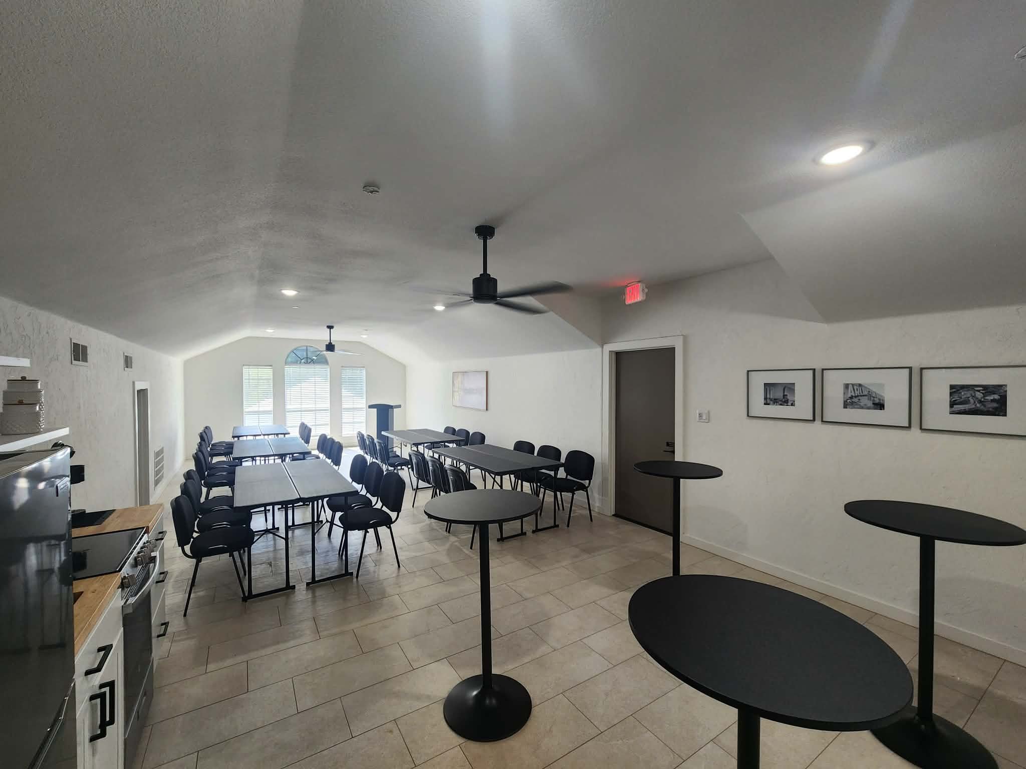 A spacious room with white walls and a high ceiling. The room has multiple tables and black chairs arranged around them. The ceiling features black ceiling fans and recessed lights. There are framed pictures on the wall, and large windows with blinds at the far end letting in natural light.