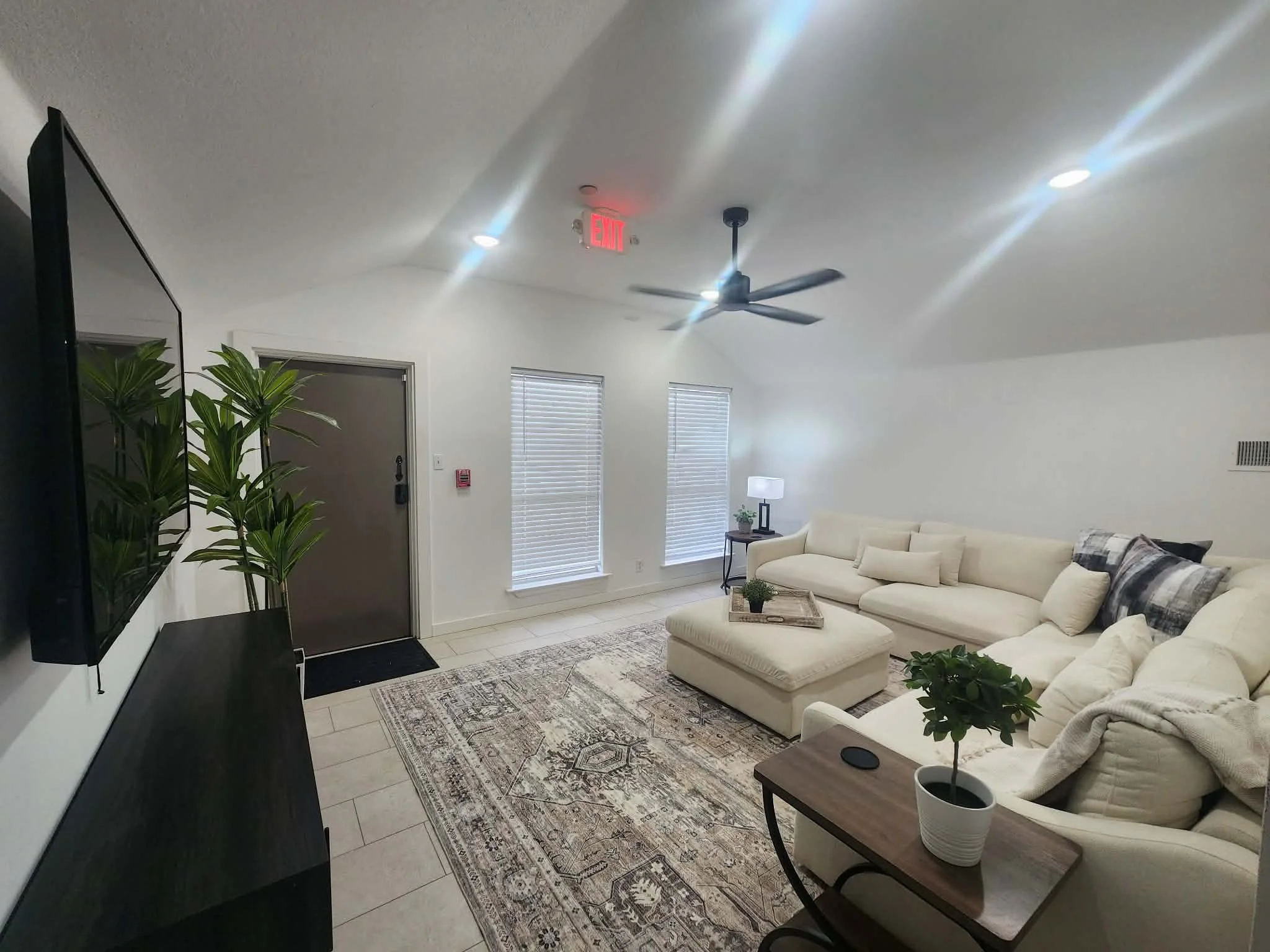 A living room with white walls and ceiling, a ceiling fan, and a large beige sectional couch with pillows. There are two windows with blinds, a small side table with a lamp, and several potted plants. A patterned rug covers the floor, and a black entertainment center or cabinet is on the left side.