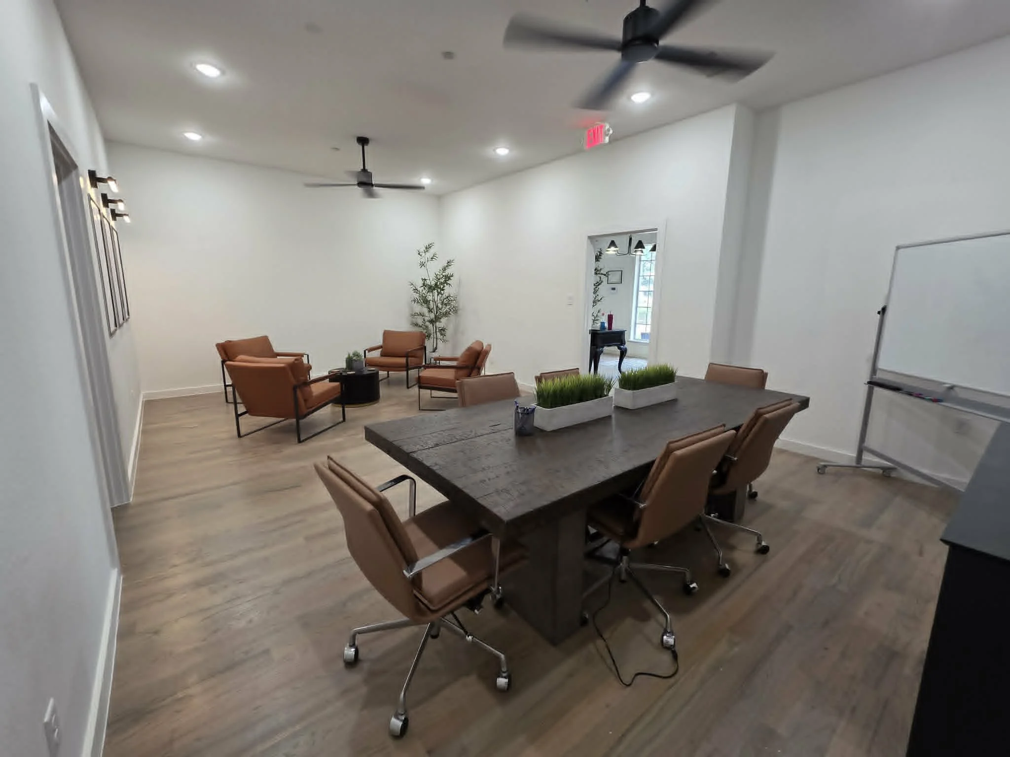 A room with a large rectangular table surrounded by tan leather rolling chairs, a whiteboard on the wall, a small side table, a potted plant, and a doorway leading to another room with a decorative table and hanging ceiling lights.