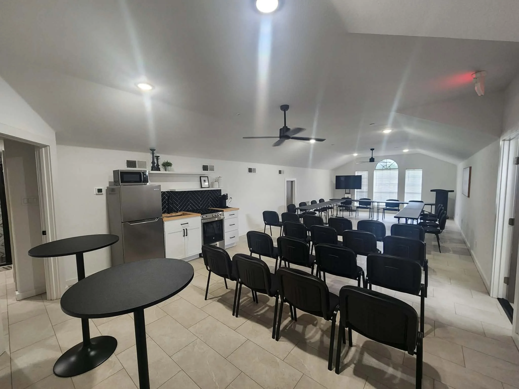 A spacious room with a kitchen area and seating arranged for a meeting or presentation. The ceiling has multiple recessed lights and a ceiling fan. There are several black chairs arranged around the room, and tables are set up for attendees. Windows with blinds let in natural light.