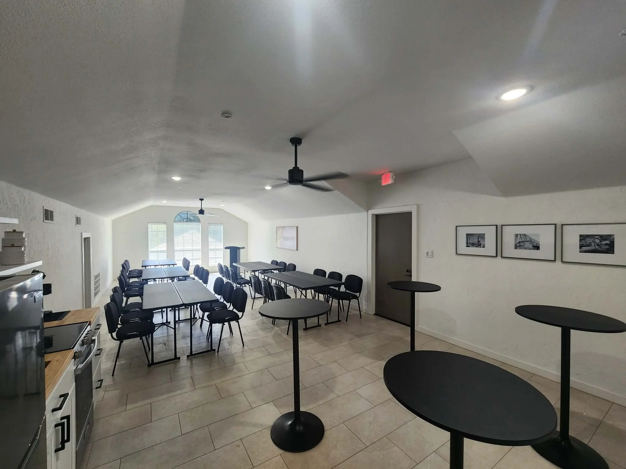 Interior of a bright room with tables and chairs arranged for a meeting or gathering, ceiling fans and lights, framed pictures on the wall, and large windows.