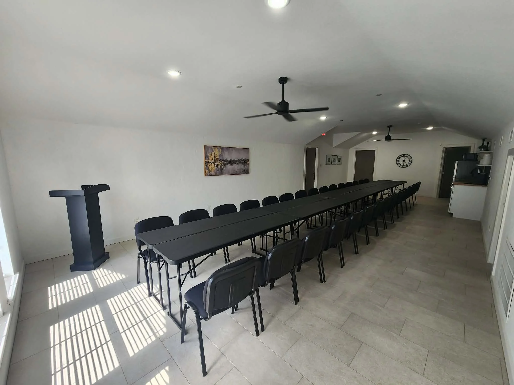 Long black conference table with black chairs in a well-lit room with white walls, ceiling fans, and artwork on the wall.
