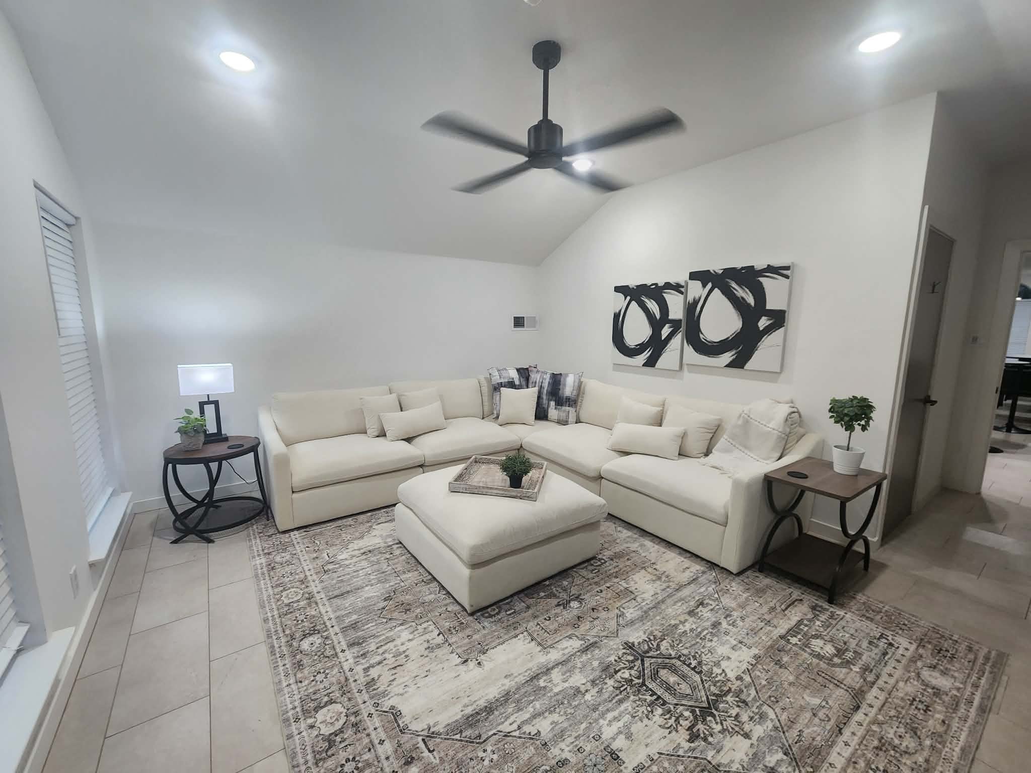 Living room with white sectional sofa, two black and white abstract paintings on the wall, a small side table with a potted plant and a lamp, a large area rug, and a ceiling fan.