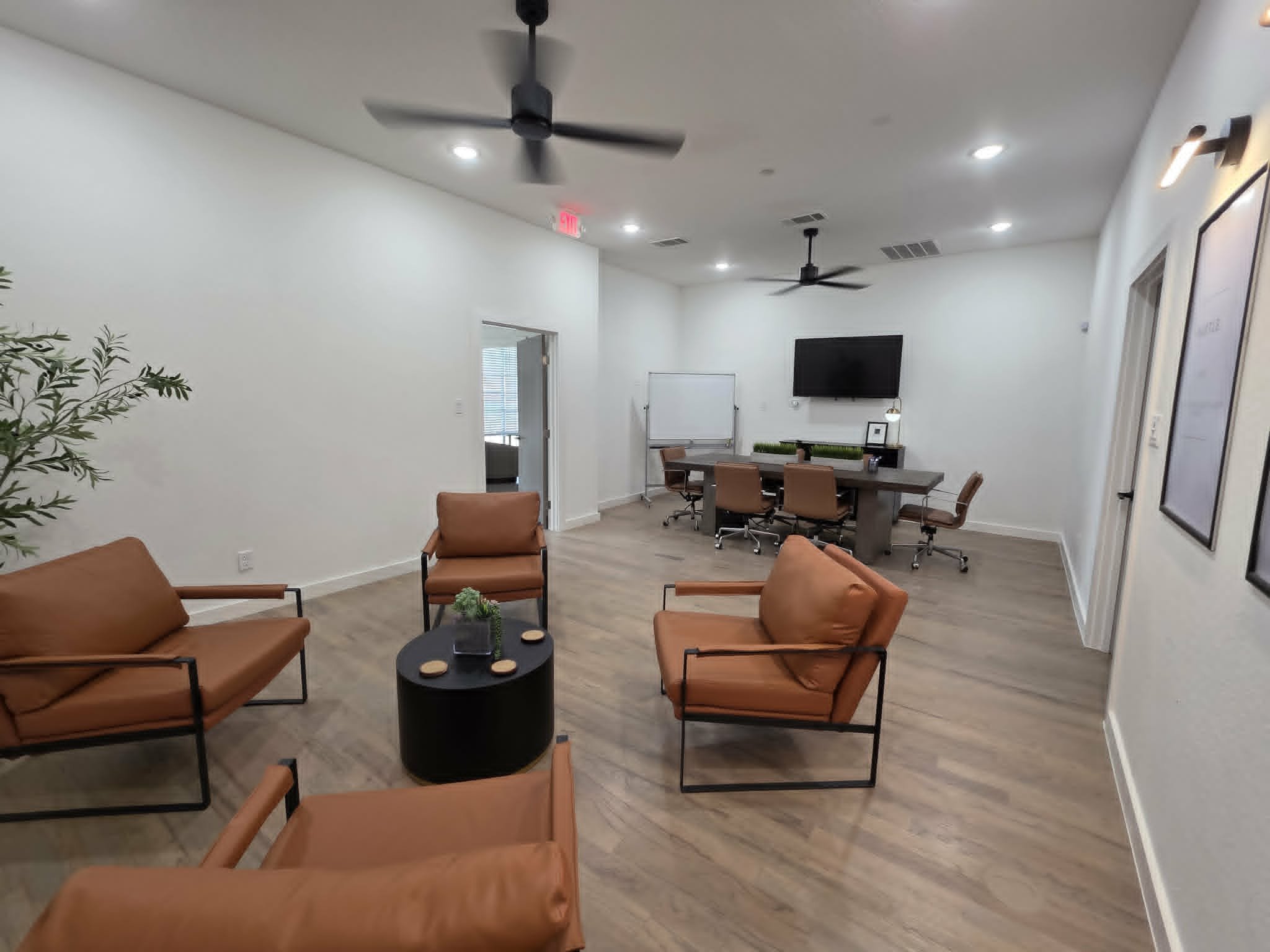Office lounge with brown chairs around a black round table, a wall-mounted TV, white walls, a white dry erase board, and ceiling fans.