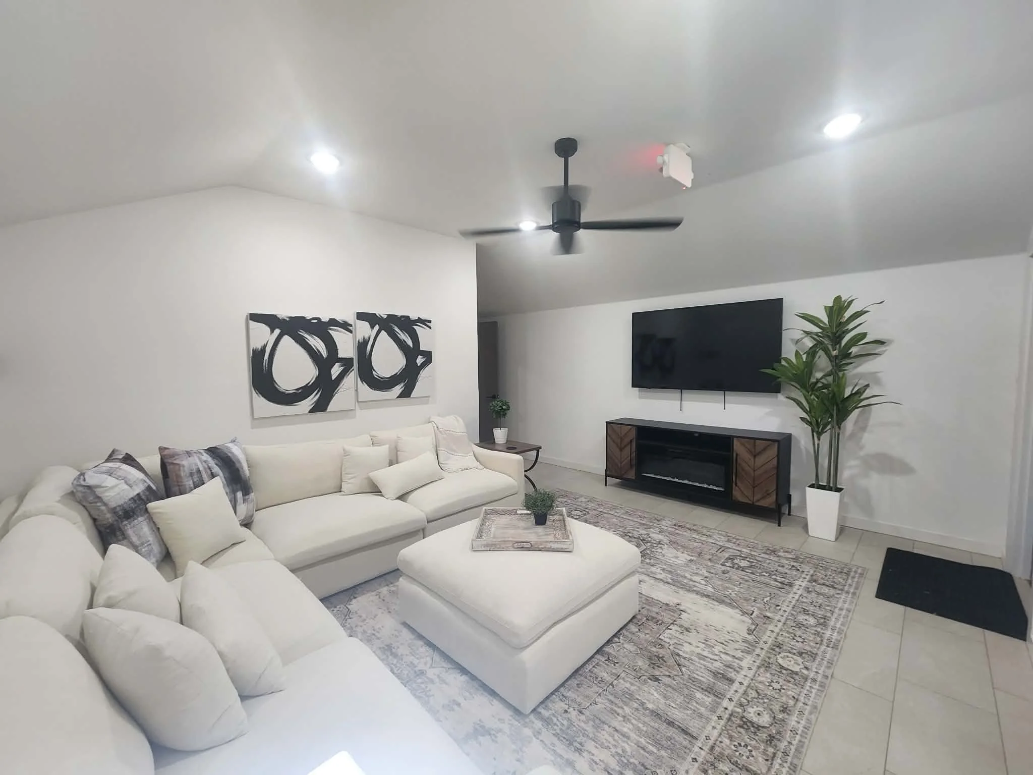 A modern living room with a large white sectional sofa adorned with plaid and neutral pillows, a patterned area rug, a wall-mounted TV, abstract black-and-white art, a wooden media console, a tall potted plant, a ceiling fan, and recessed lighting.
