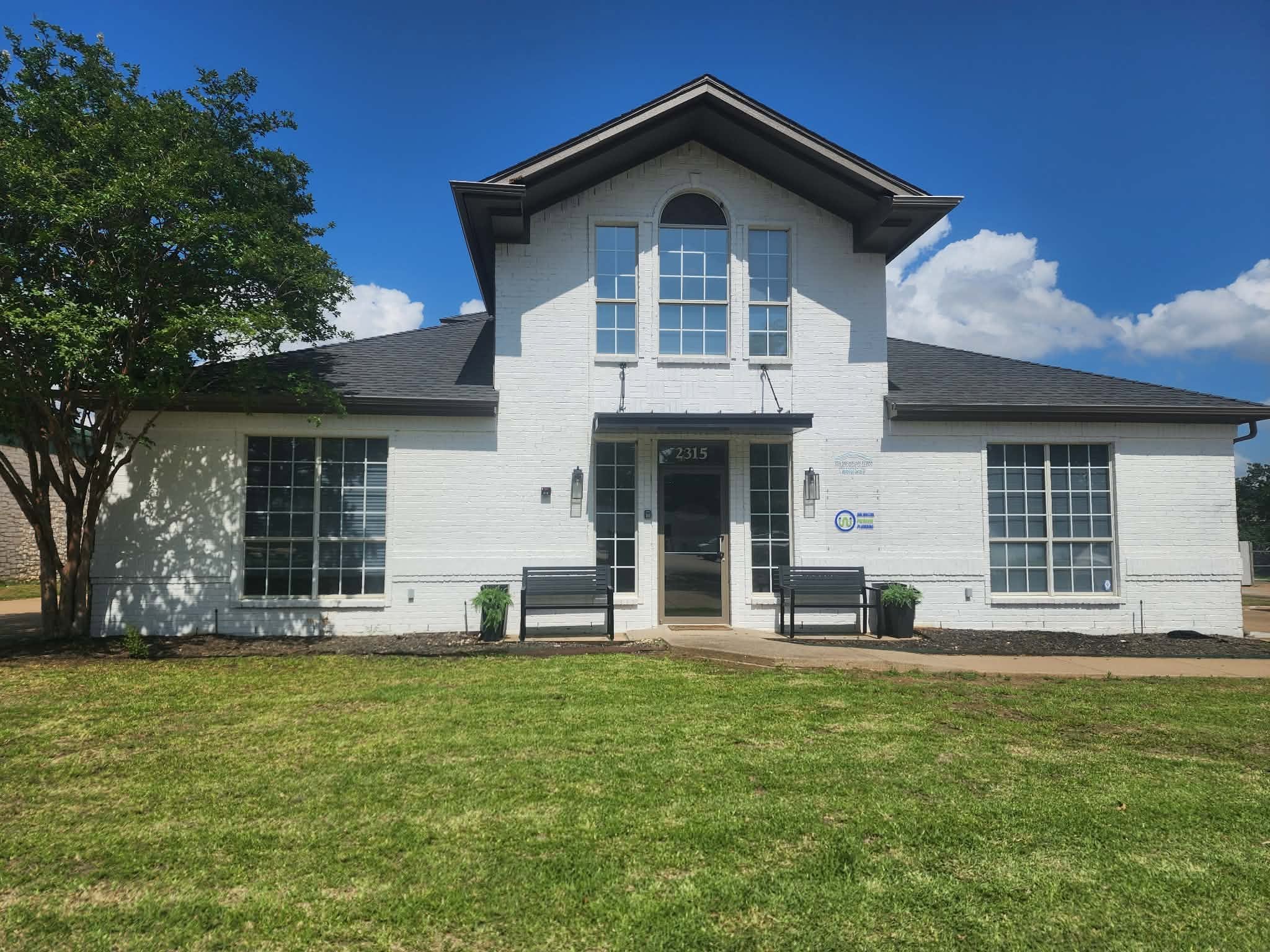 A white two-story house with large front windows, a black door, and a gabled roof, surrounded by a green lawn and a tree on the left, under a clear blue sky with some white clouds.