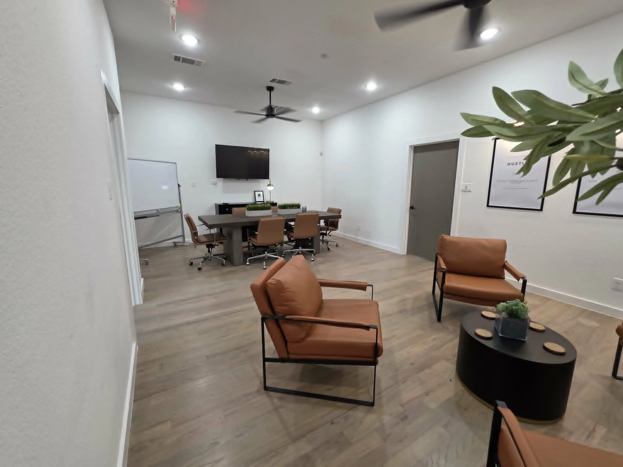 Modern conference room with a large wooden table surrounded by brown chairs, wall-mounted TV, whiteboard, ceiling fan, and wall art. Furniture includes a lounge chair and round side table with decorative items.