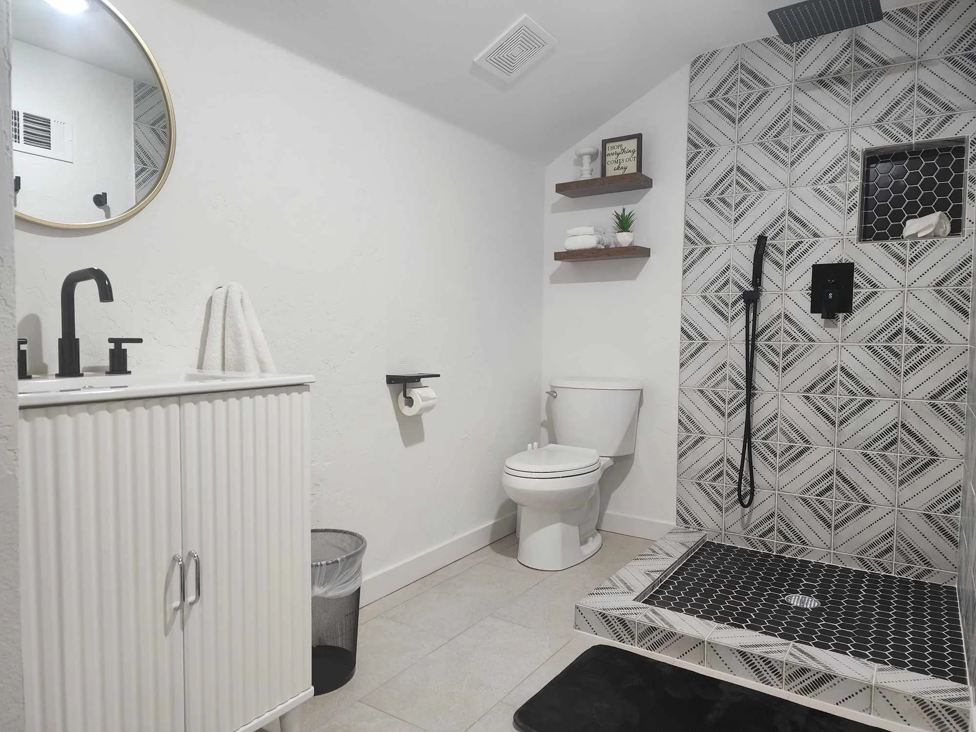 A modern bathroom with a black and white color scheme. Features include a round mirror, black faucets, a white shelf with towels, a white toilet, a small potted plant, and a geometric-patterned shower area with black tile accents.