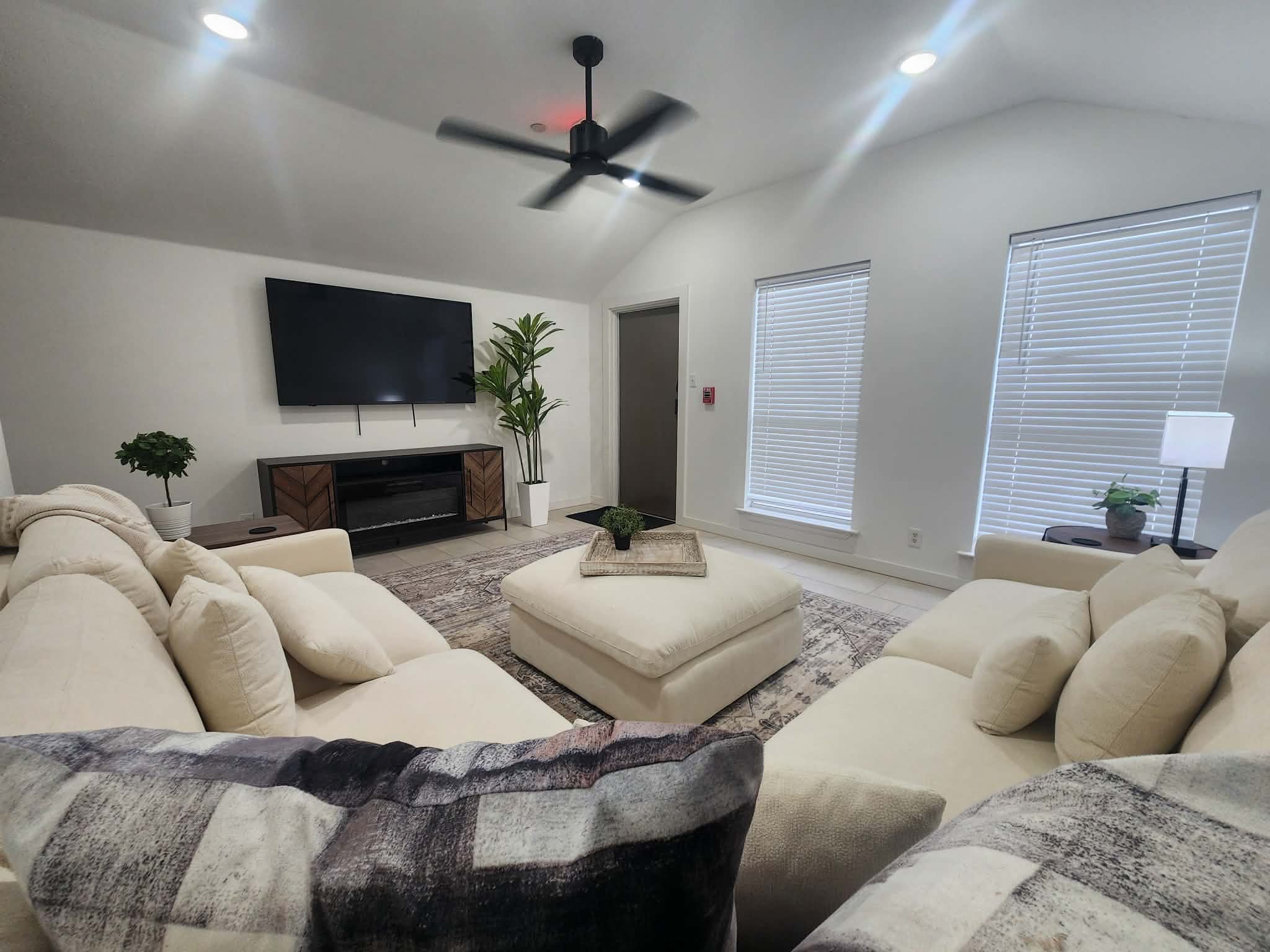 Living room with cream-colored sectional sofa, black and white rug, television, tall indoor plants, white blinds on windows, ceiling fan, and white walls.