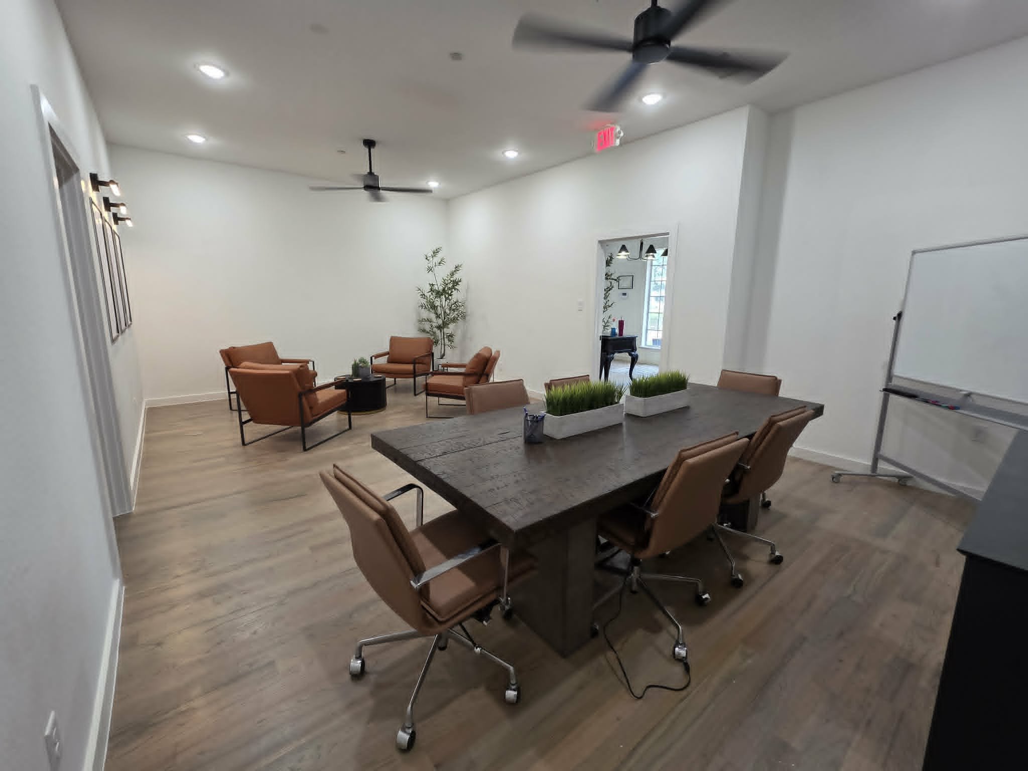 A modern conference room featuring a large wooden table surrounded by twelve tan leather chairs on wheels. There are decorative planters with green grass on the table. The room has white walls, wooden flooring, a ceiling fan, and recessed lighting. In the corner, there is a whiteboard, and in the background, a small sitting area with two chairs, a small table, a potted plant, and a window with vertical blinds.