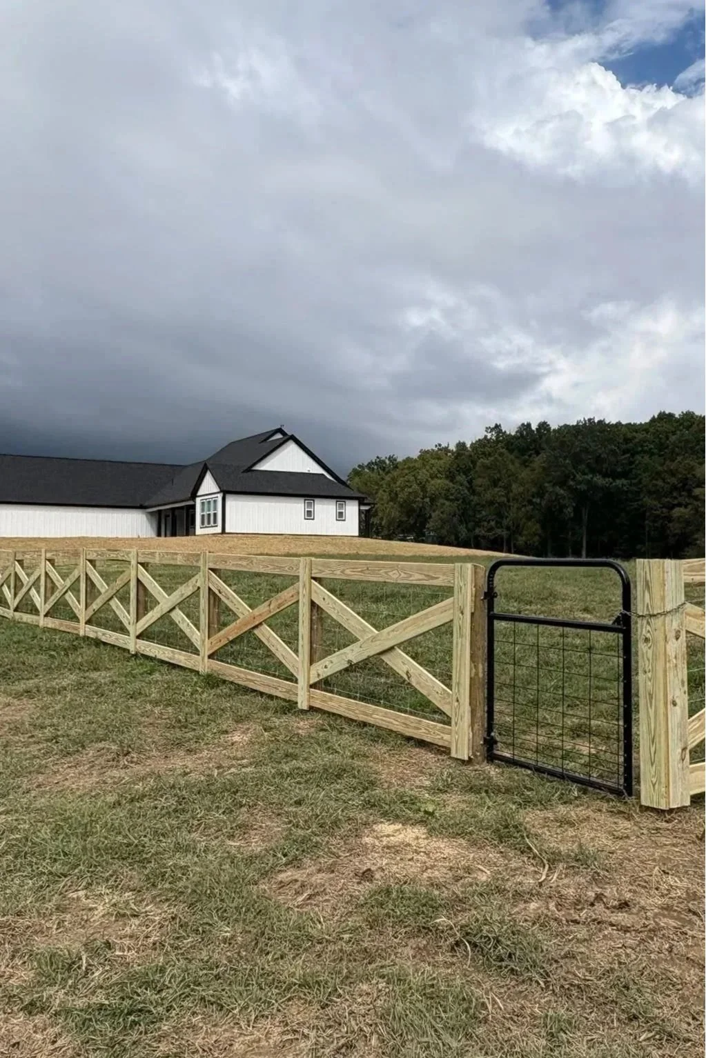 Custom treated wood cross-buck farm fence with wire mesh backing and a black metal pasture gate installed by Lane's Fence, leading up a grassy hill toward a modern white farmhouse.