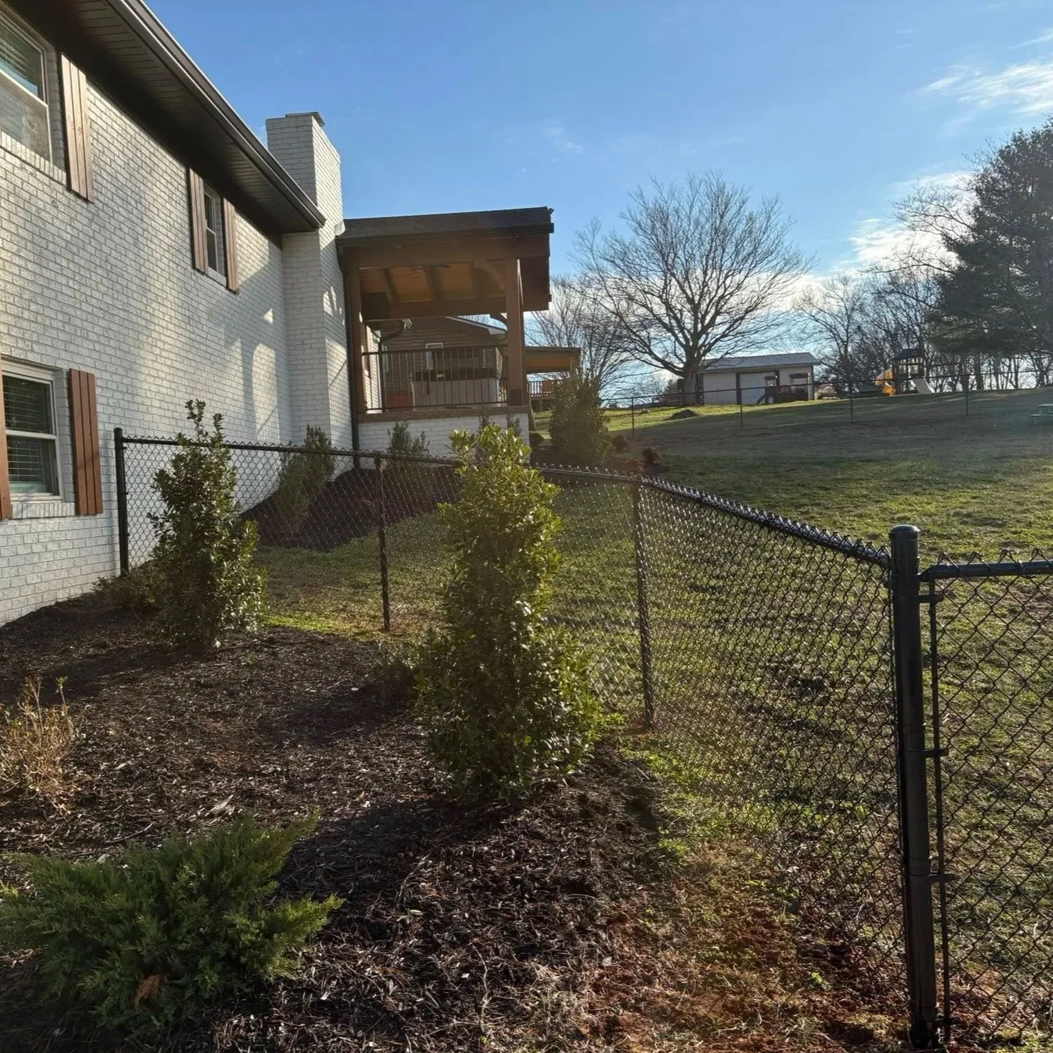 Black vinyl-coated chain-link fence installed by Lane's Fence, bordering a landscaped residential yard and seamlessly following the slope away from a white brick home.