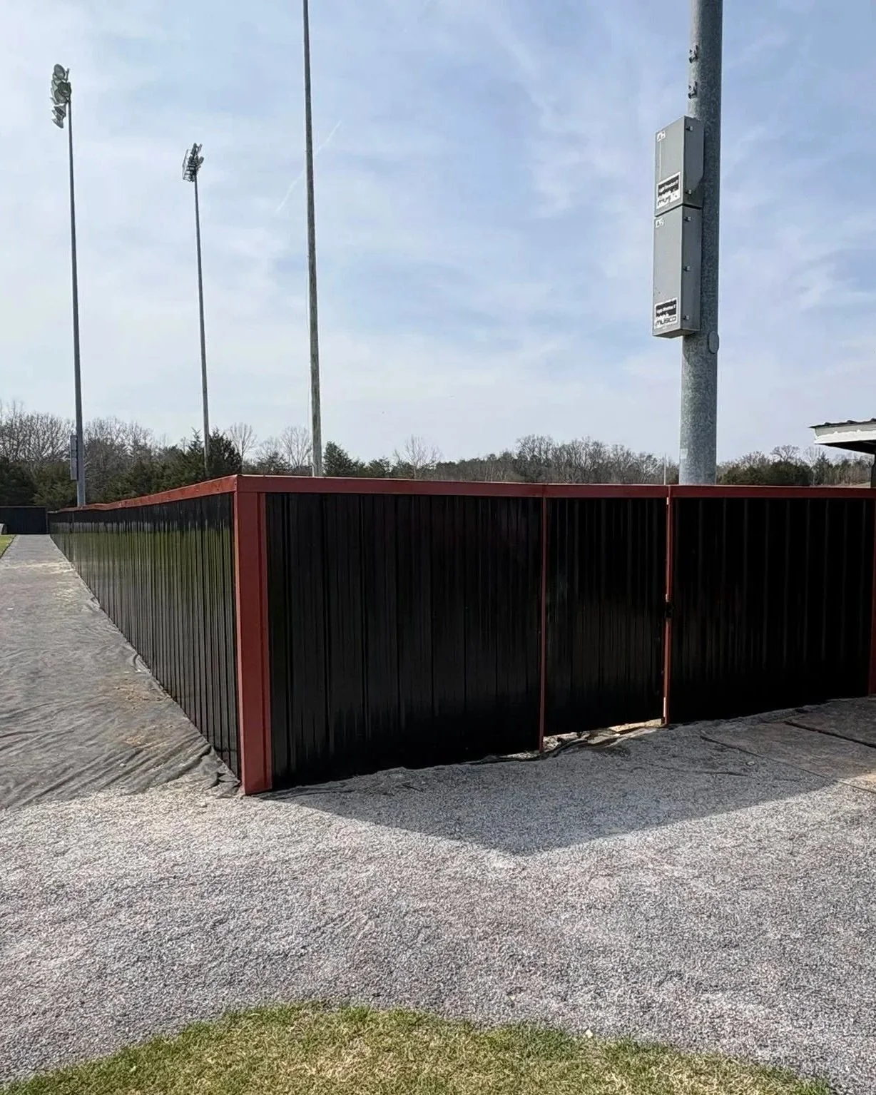 Solid black metal panel athletic field fence with a red top rail and built-in access gate, installed near a sports facility dugout by Lane's Fence.