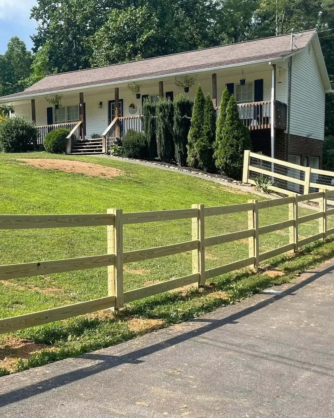 Newly installed treated wood post-and-rail fence with wire mesh backing by Lane's Fence, bordering a sloped residential front yard and asphalt driveway.