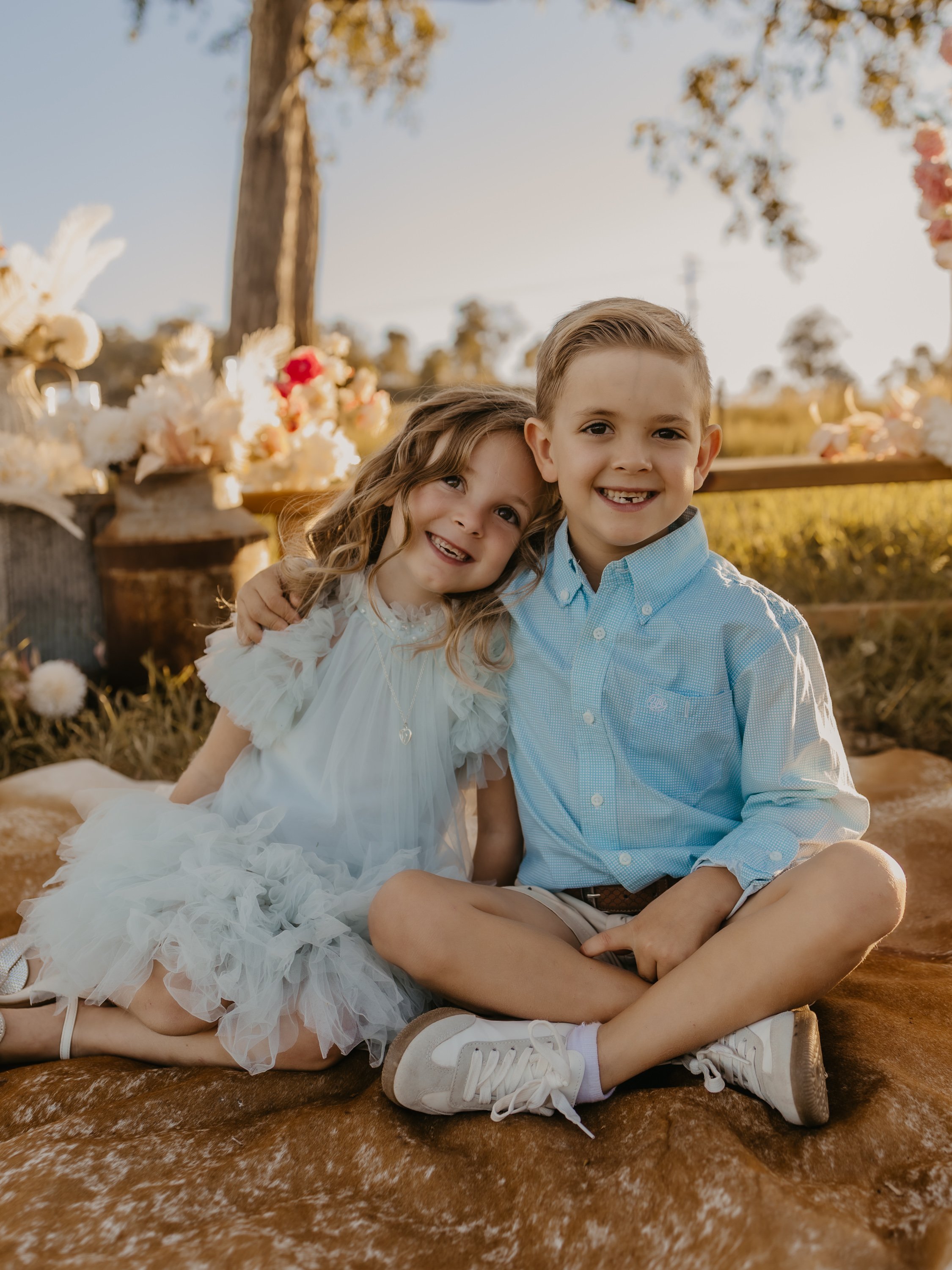 The COWGRL Edit | Family Session - A boy and girl sitting on a blanket outdoors during sunset, smiling and posing for the camera, with a background of trees, a wooden table with flower arrangements, and a clear sky. The Cowgirl Edit