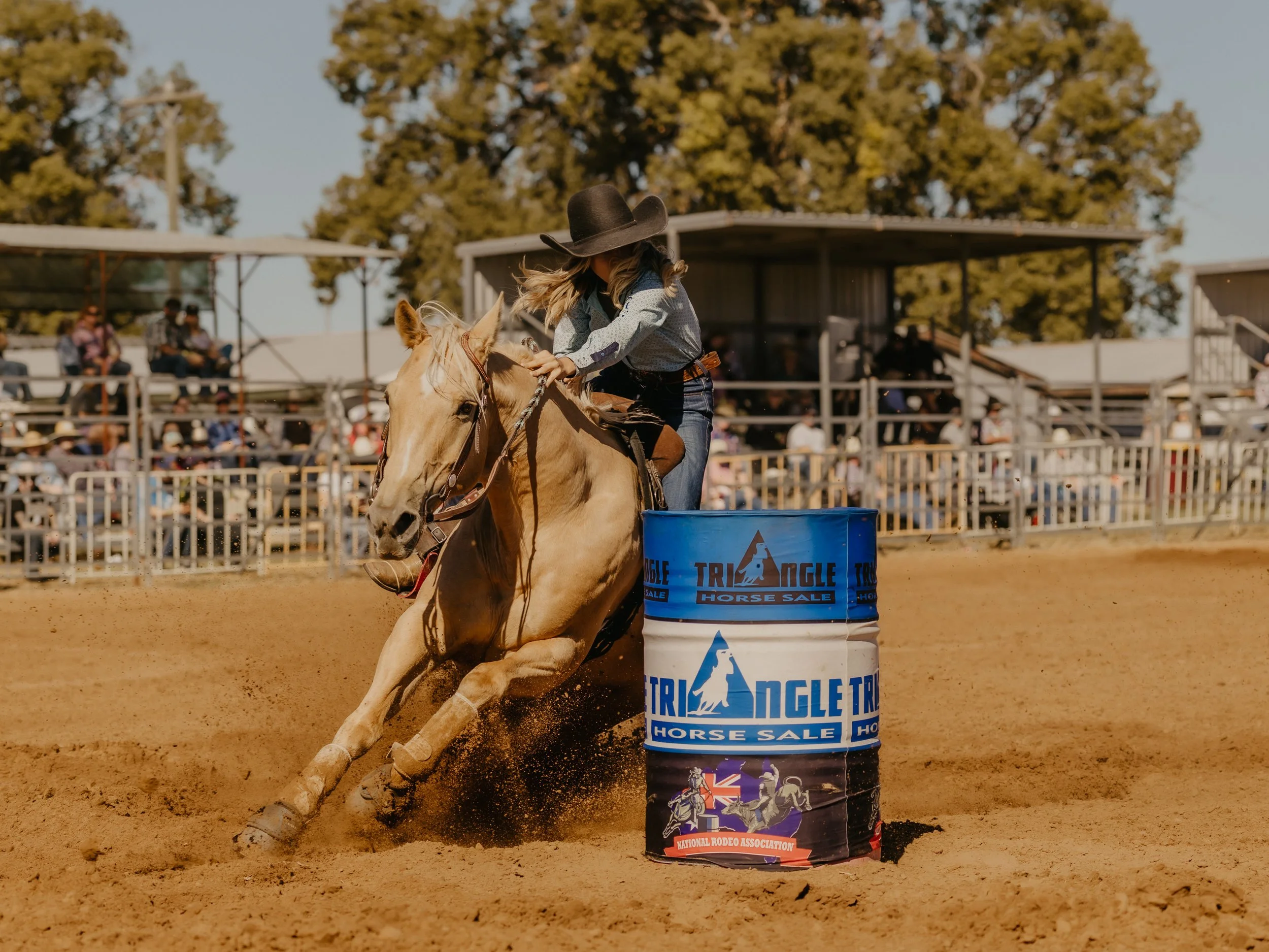 The WESTRN Edit | Western Sports Photography - A woman wearing a cowboy hat and jeans riding a light-colored horse around a barrel at a rodeo event, with spectators in the background and trees in the distance. The Western Edit