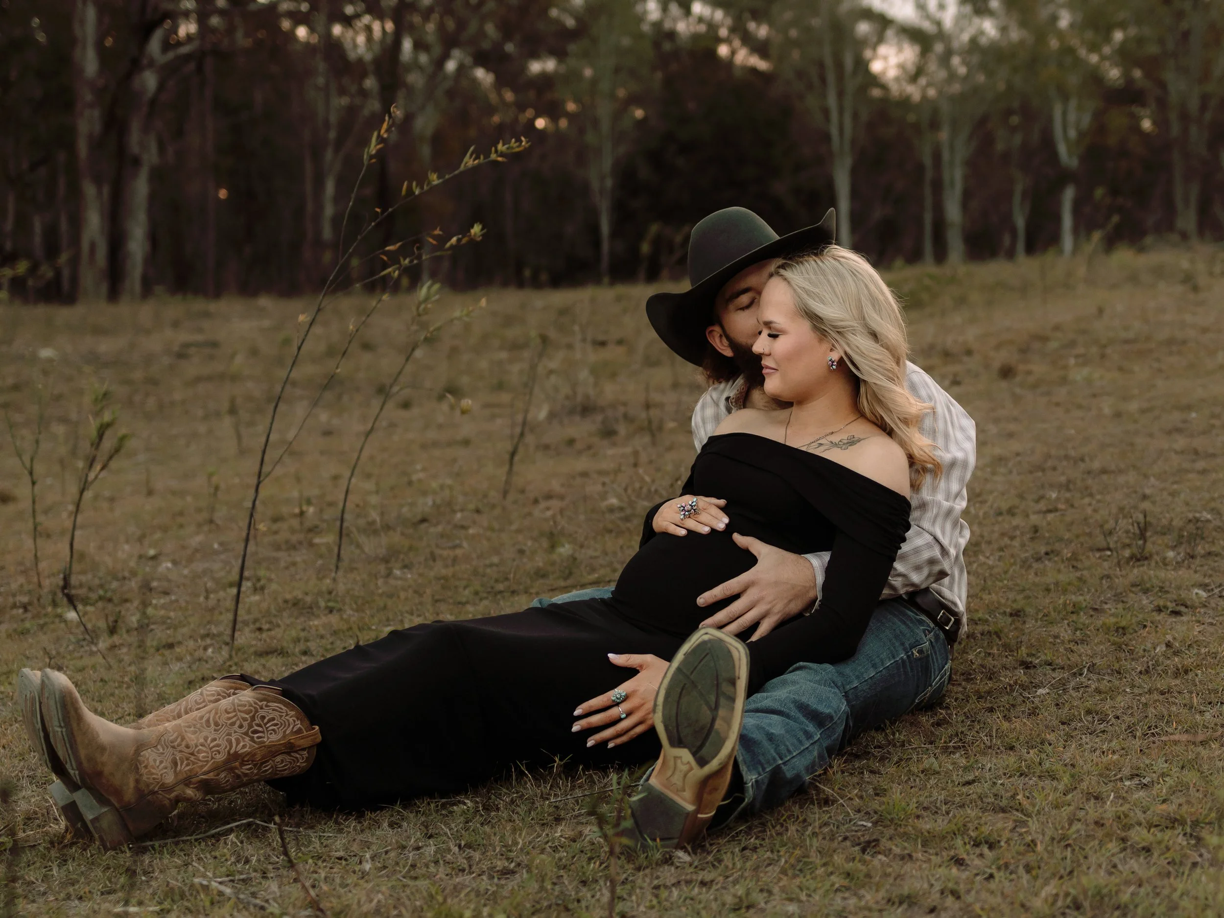 A couple sitting on the grass in a field, the pregnant woman is smiling and embracing her partner, while he kisses her forehead; they are surrounded by trees in the background, during sunset.