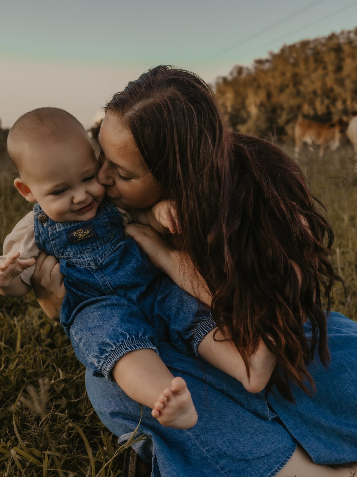 The COWGRL Edit | Motherhood Session - A woman with long dark hair sitting on grass, holding a smiling toddler, in a field with trees and horses in the background.
