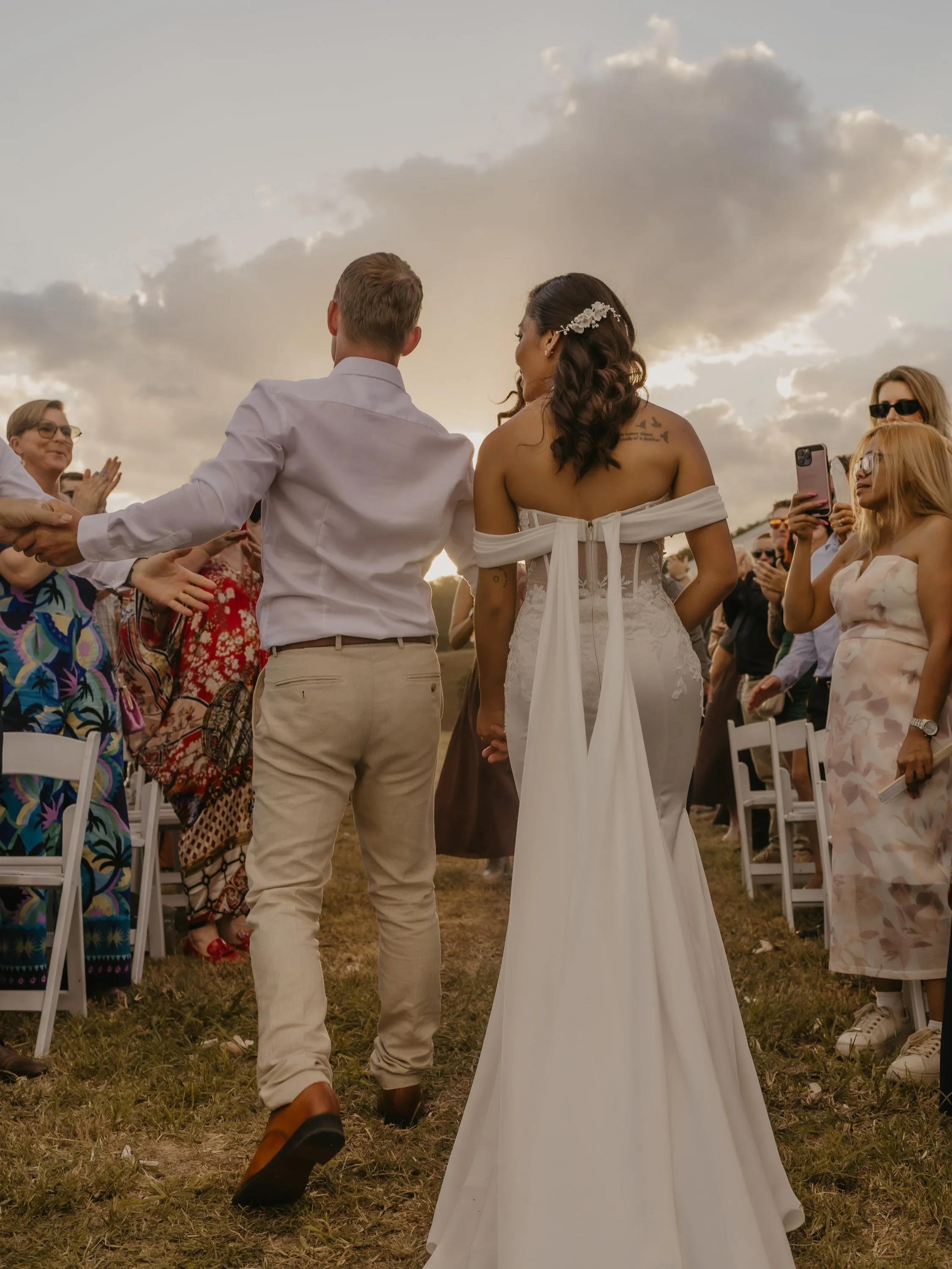 The COWGRL Edit | Wedding Package - A couple dressed in wedding attire holding hands and walking down the aisle during an outdoor wedding ceremony at sunset, surrounded by friends and family.
