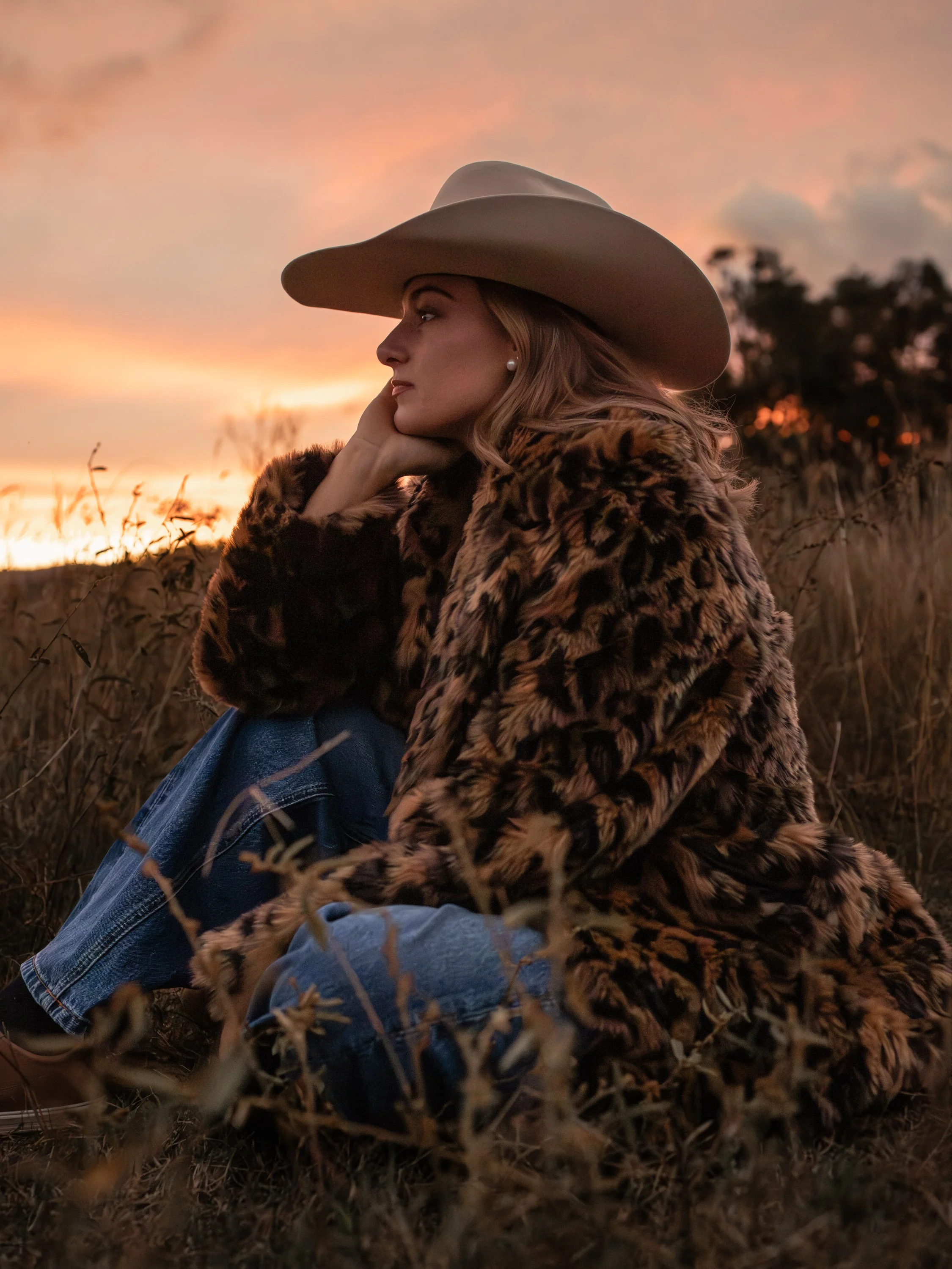 The COWGRL Edit | Empowerment Session - A woman wearing a large beige cowboy hat, pearl earrings, a leopard print coat, and blue jeans sitting in a field during sunset, with her hand resting on her face and looking to the side. The Cowgirl Edit