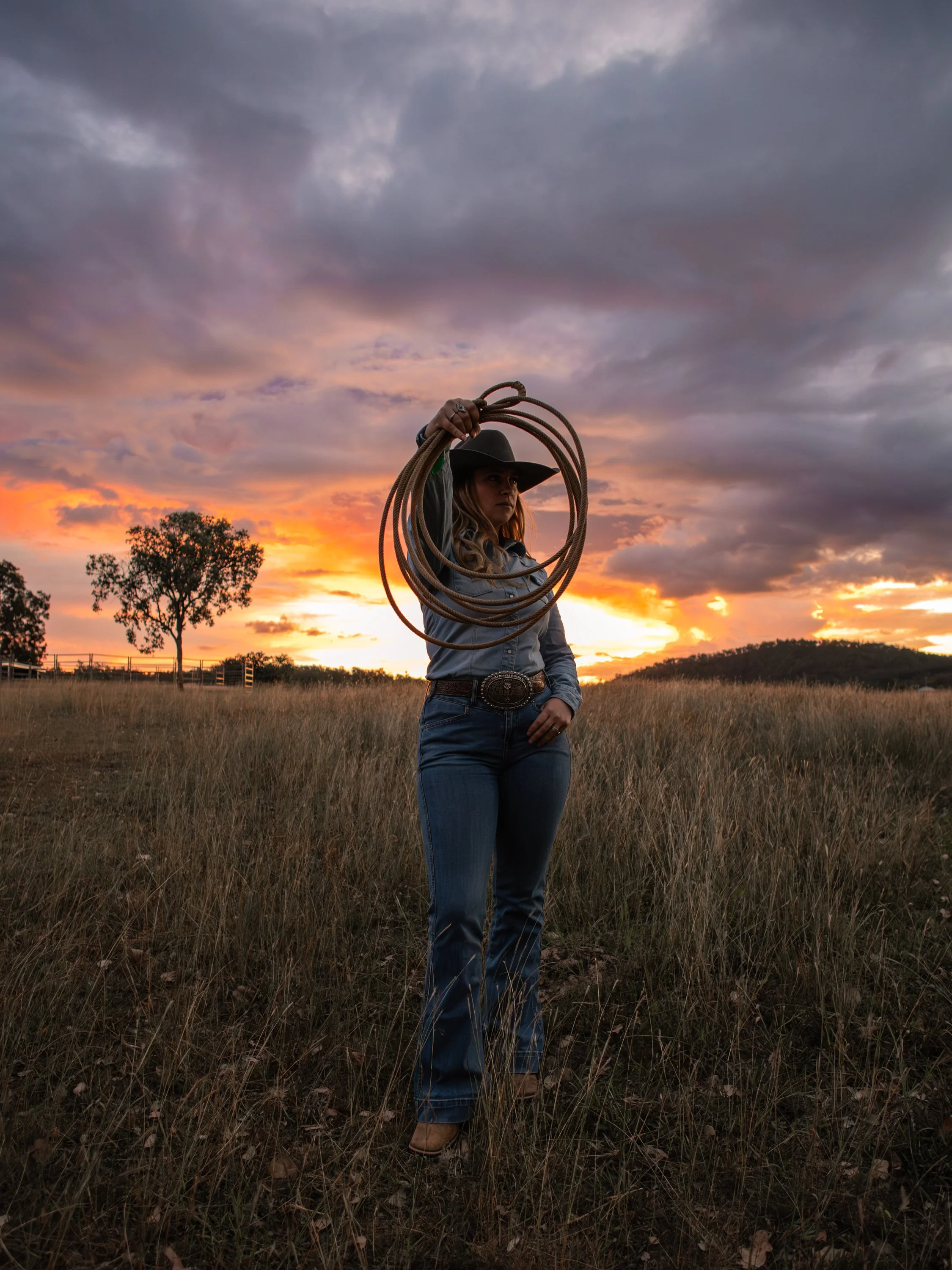 The COWGRL Edit | Empowerment Session - A woman holding a lasso stands in a grassy field at sunset, wearing a cowboy hat and jeans, with a sky filled with clouds and trees in the background. The Cowgirl Edit