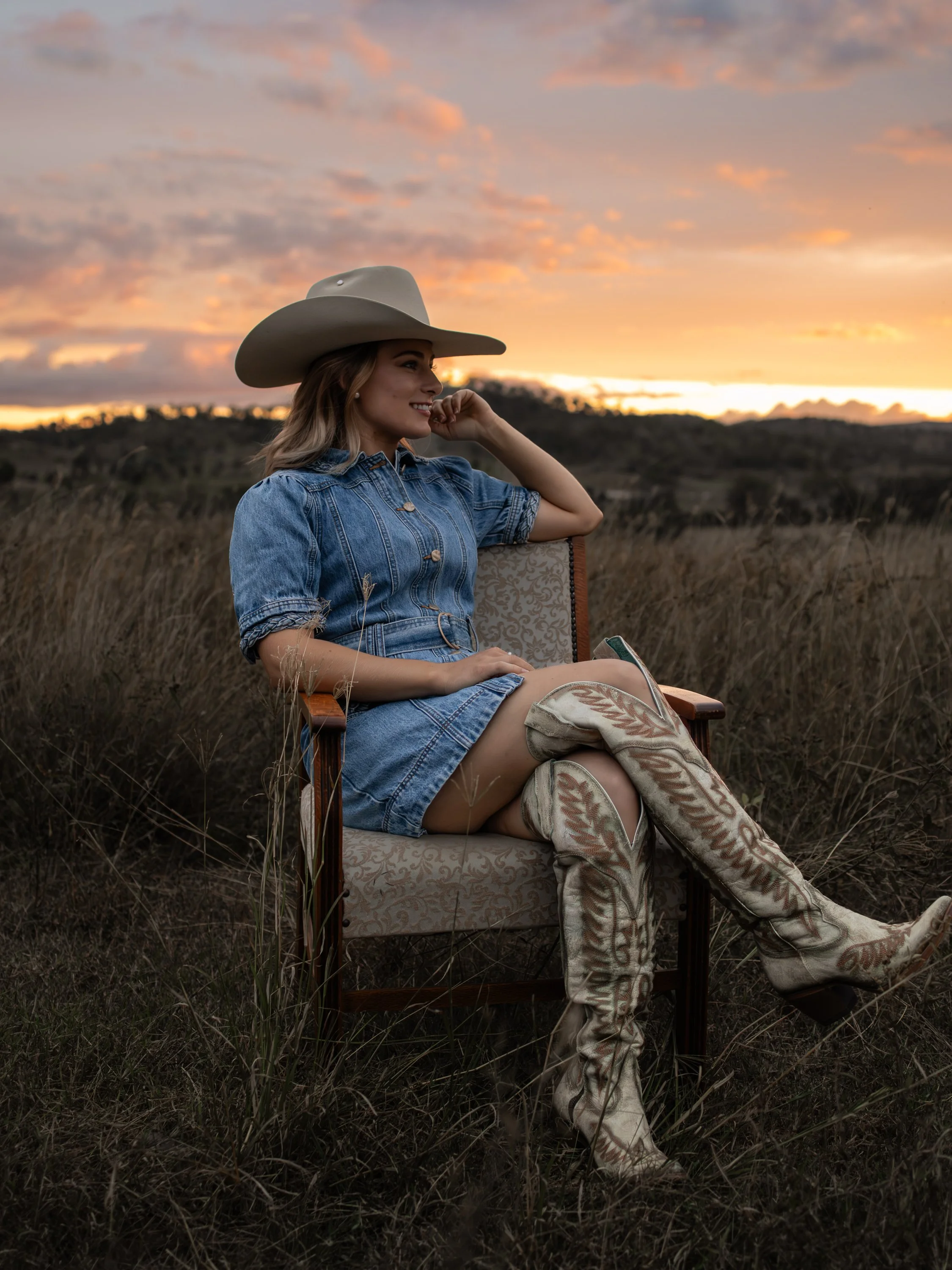 The COWGRL Edit | Empowerment Session - A woman sitting on a vintage wooden chair outdoors at sunset, wearing a denim dress, cowboy hat, and cowboy boots, in a field with tall grass. The Cowgirl Edit